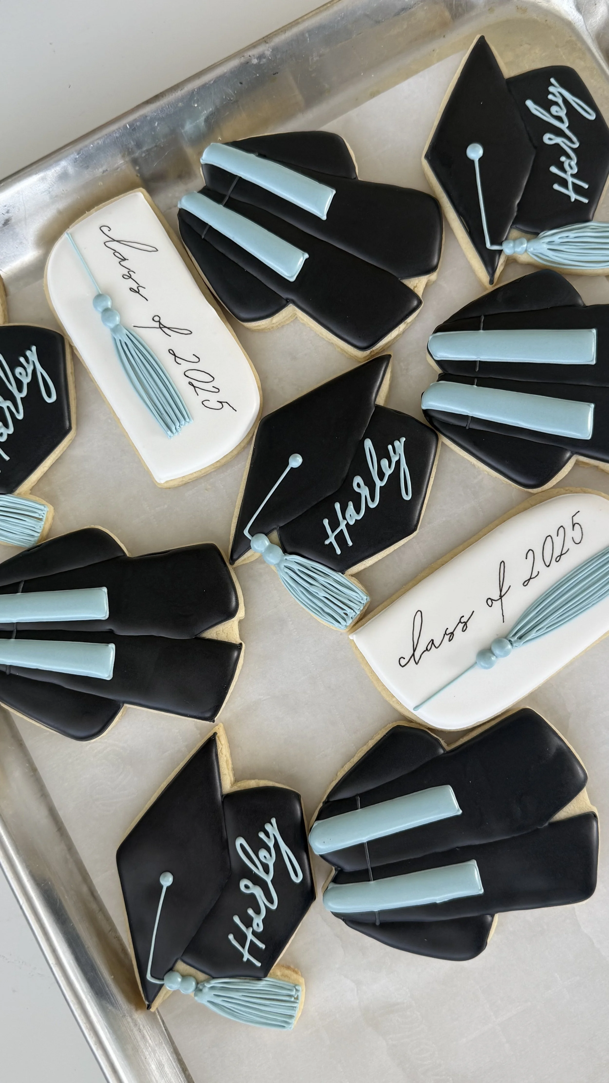 Graduation-themed cookies decorated with black, white, and blue icing, some shaped like graduation caps, and others with the words 'class of 2023' written on them.