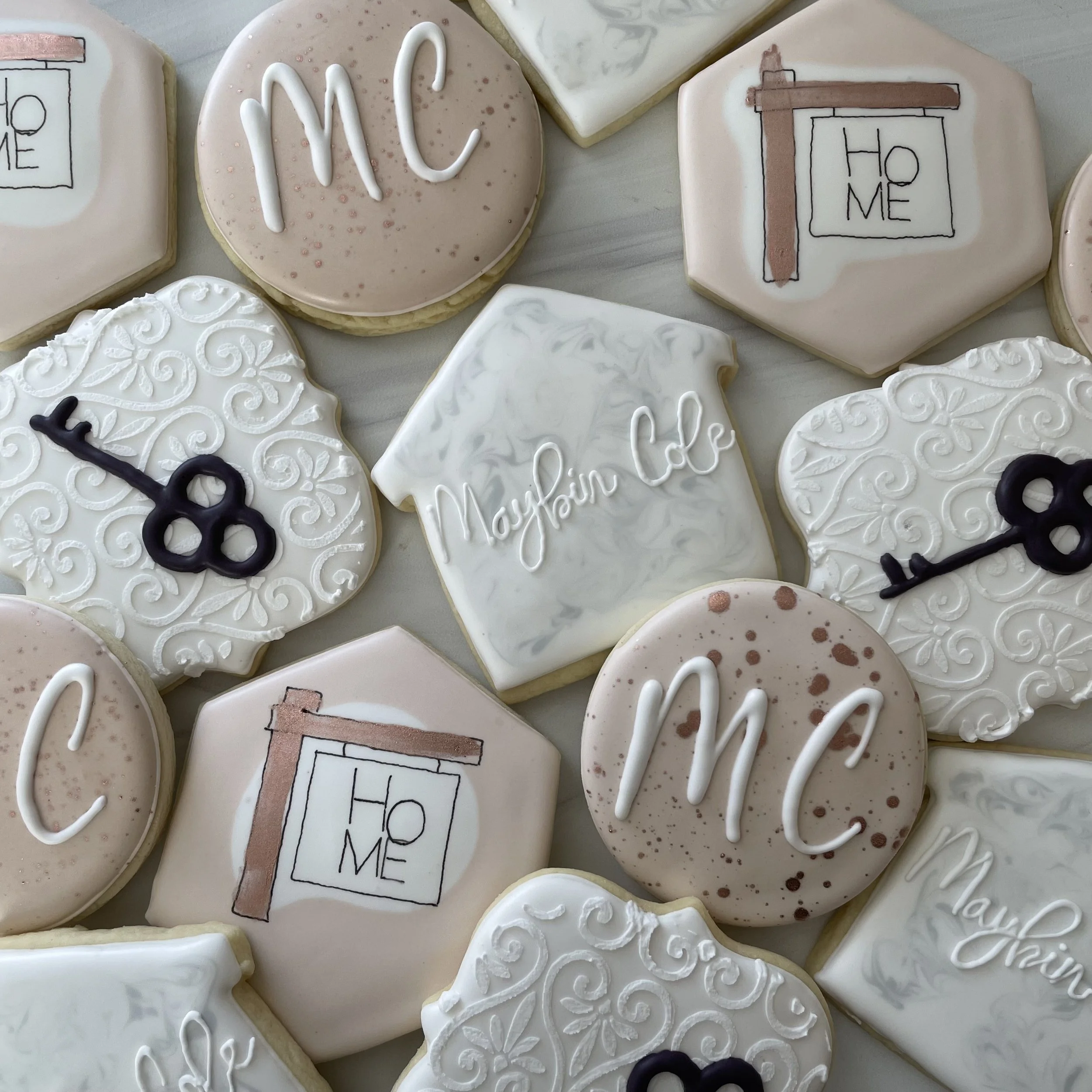 Decorated cookies with white, pink, and black icing; some cookies have 'Home' written with piping, others have intricate white icing patterns or designs, and some display words like 'My' in white icing. The cookies are hexagonal, square, or round in 