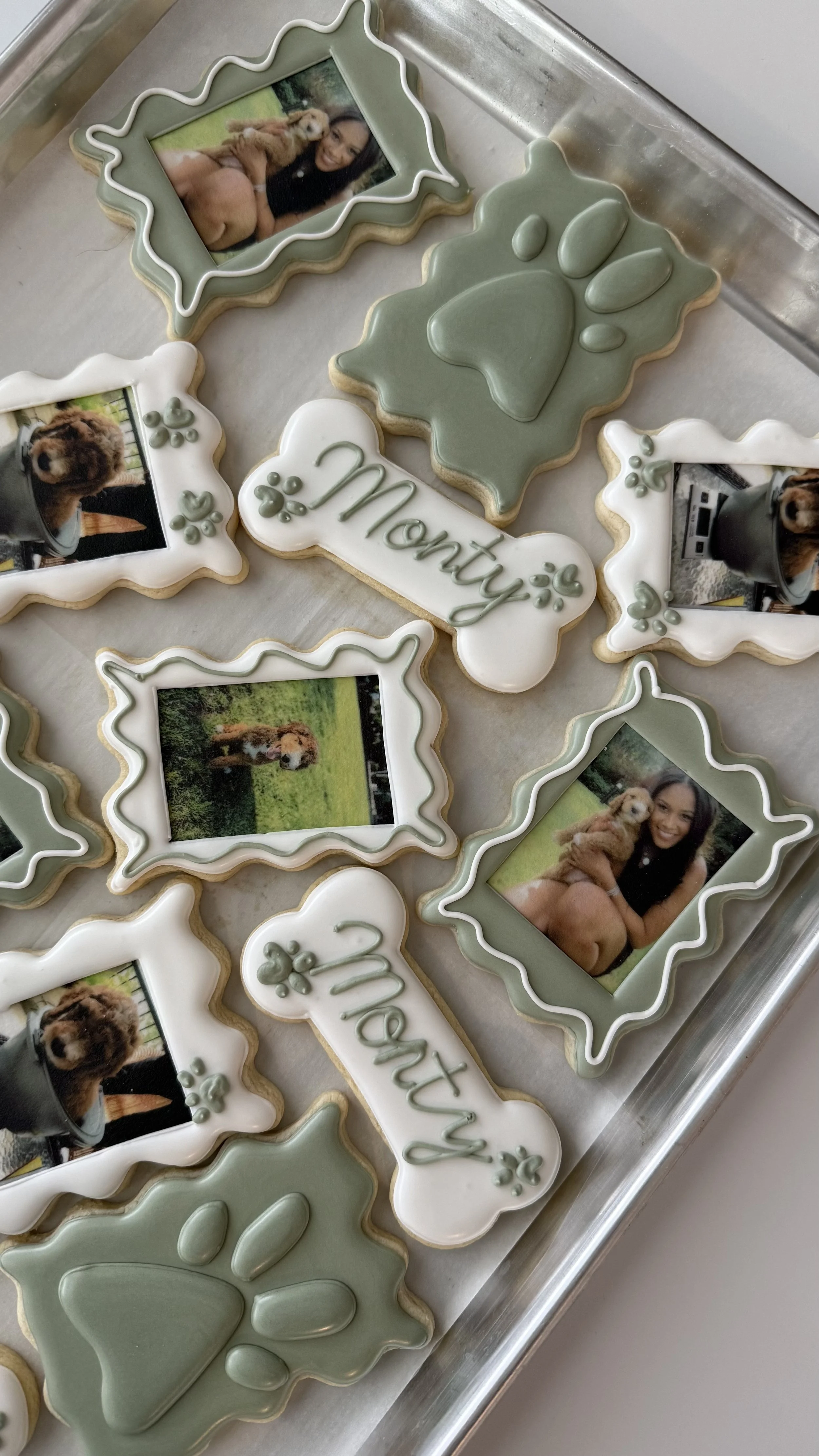 Cookies decorated with photos of a woman holding a puppy, paw prints, and the word 'Monty' in green icing, arranged on a metal tray.