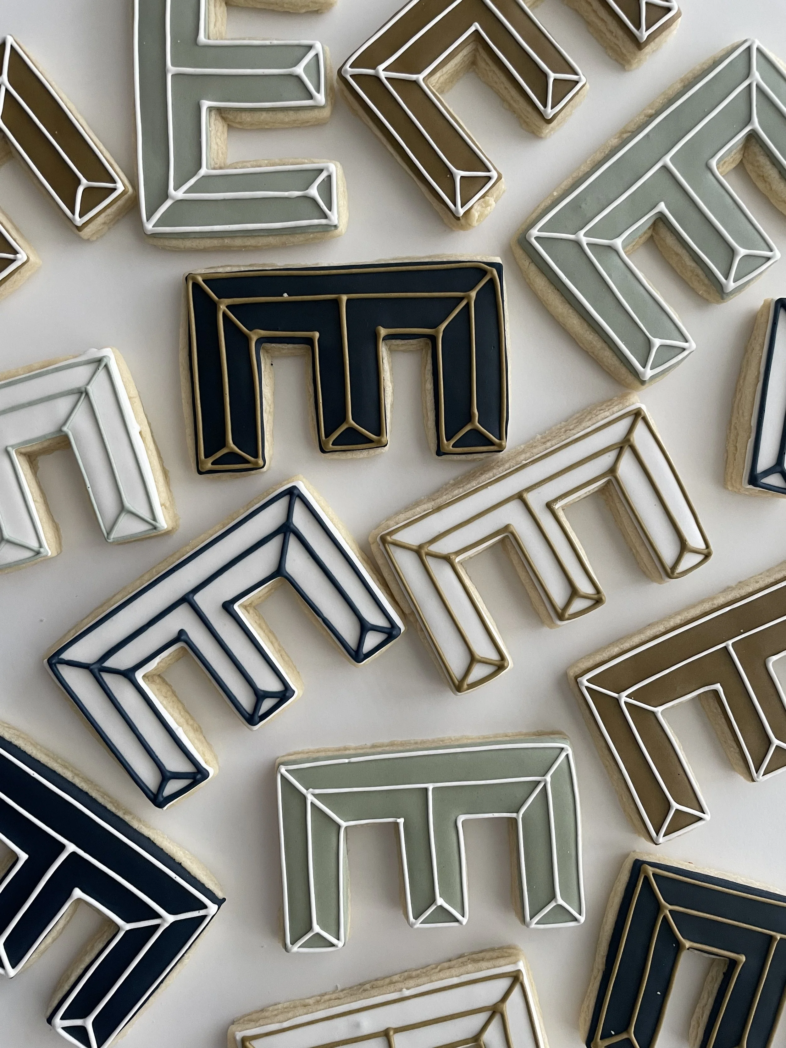 Decorative cookies shaped like the letter 'E' with geometric line designs, in various colors including brown, green, white, navy blue, and gold, laid out on a white surface.