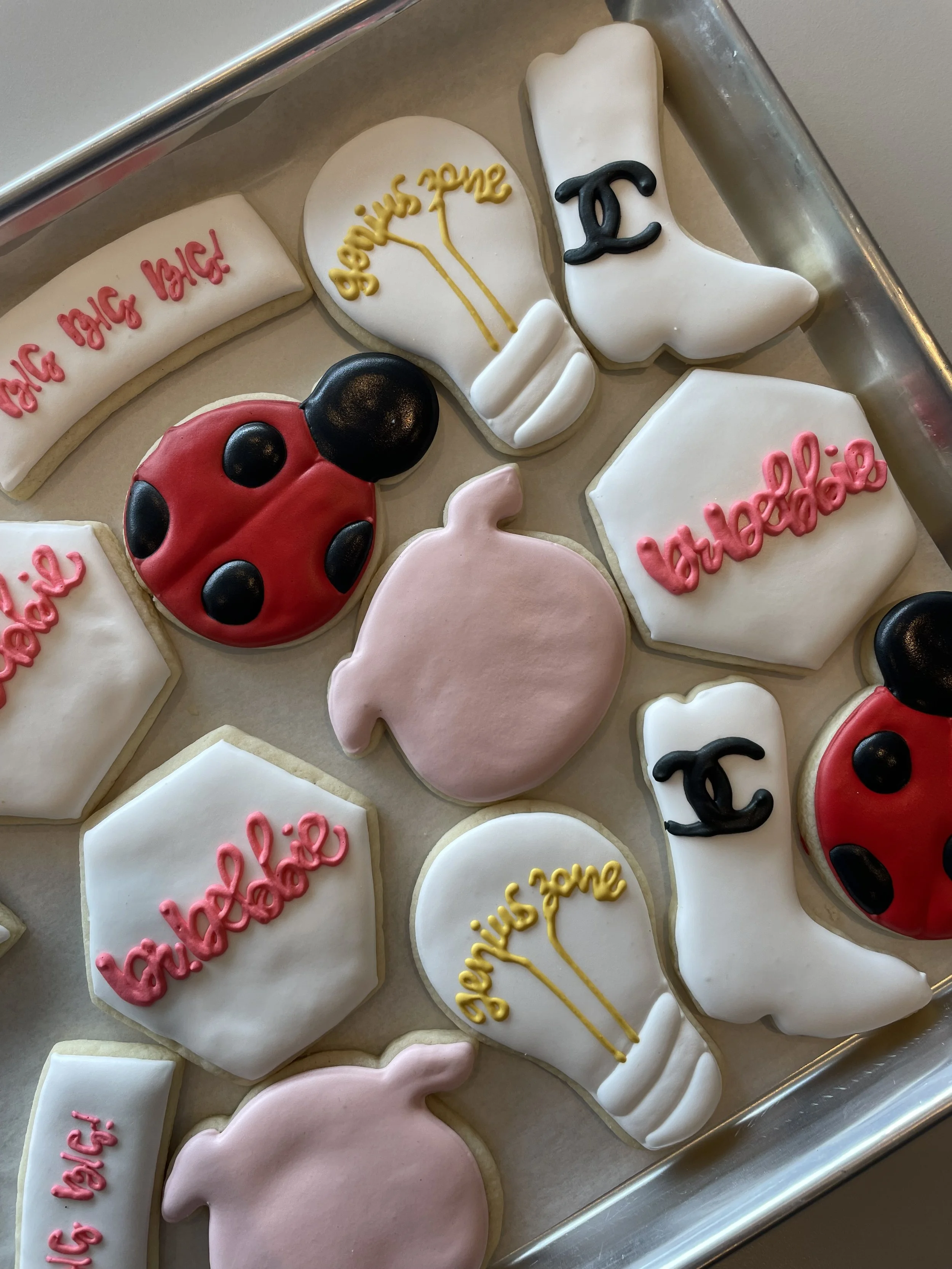 A tray of decorated sugar cookies with various shapes and messages, including red ladybug, a pink pig, white moon, and cookies with the words 'happy birthday!' and yellow speech bubble with 'say so' written on it.