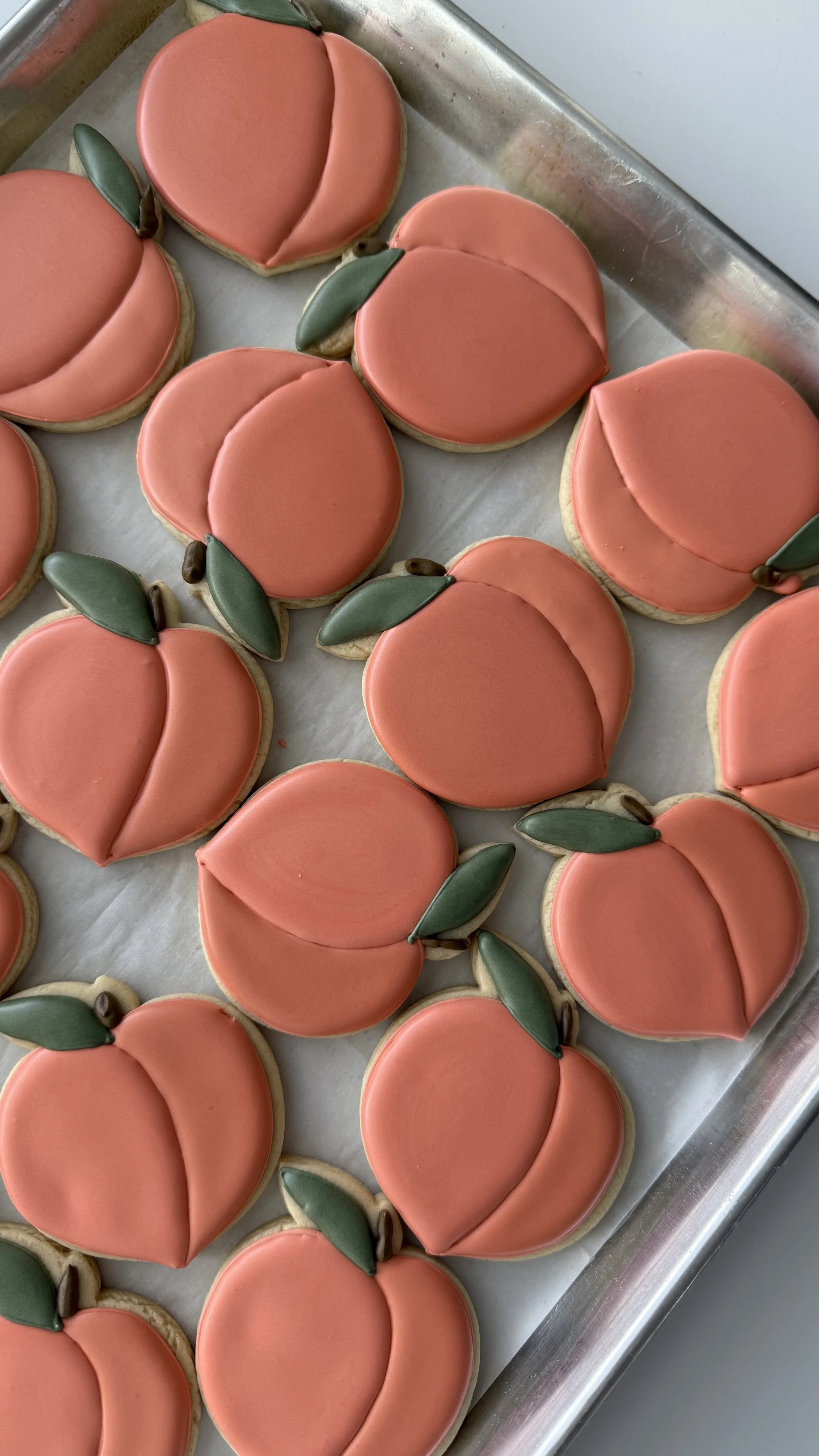 Decorated cookies shaped and decorated like peaches, with green leaves and brown stems, on a baking sheet.