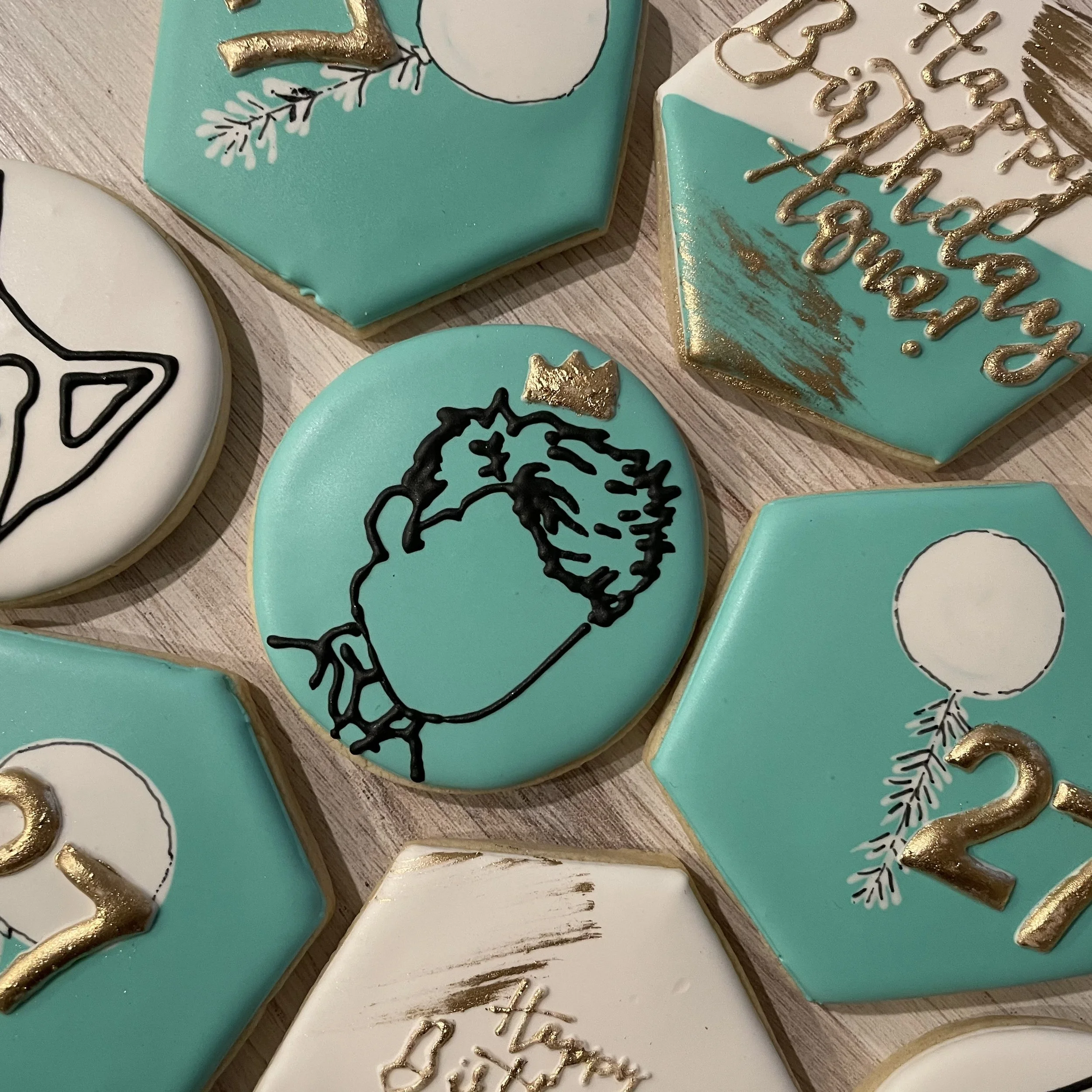 Close-up of decorated cookies with teal, white, and black icing, featuring unicorn, heart, and holiday-themed designs with gold accents.