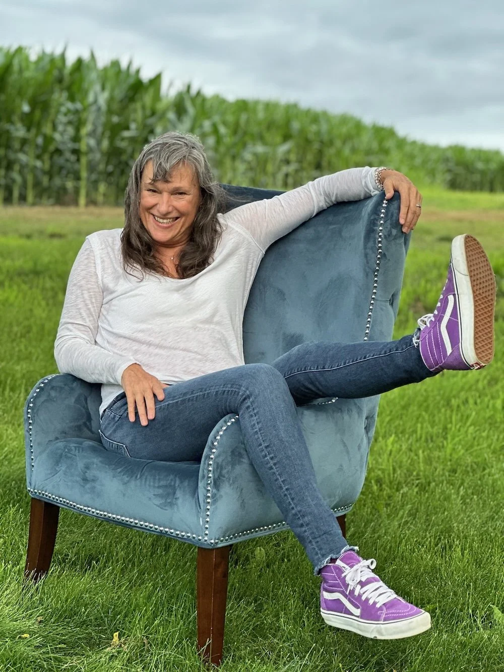 Rita Henry sitting sideways on a blue velvet armchair outdoors in a grassy field with a cornfield in the background. She is smiling, wearing a white long-sleeve shirt, jeans, and purple sneakers, with one leg resting on the armrest.