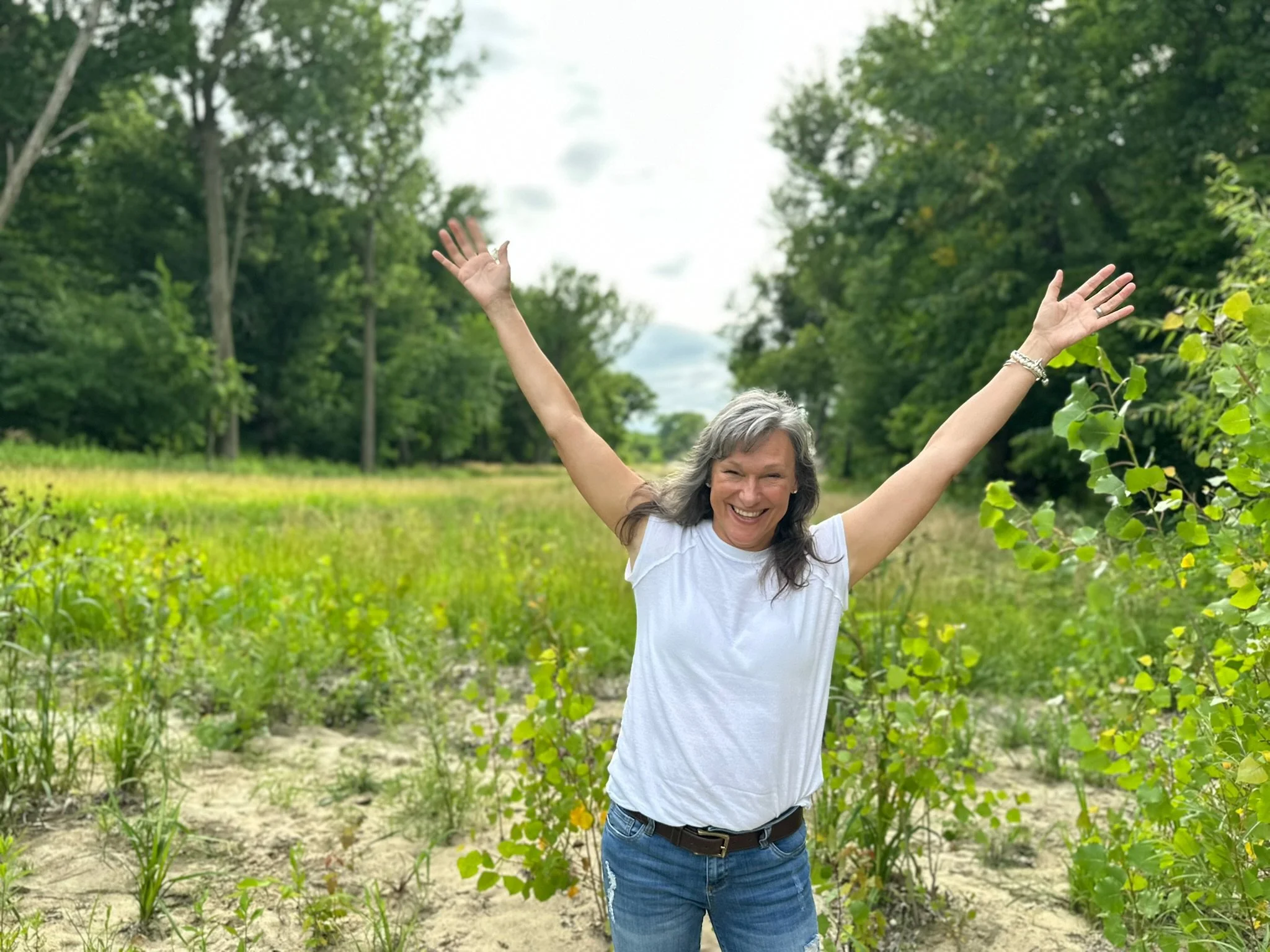 A smiling woman with gray hair raises her arms in a field with green plants, surrounded by trees, under a cloudy sky.