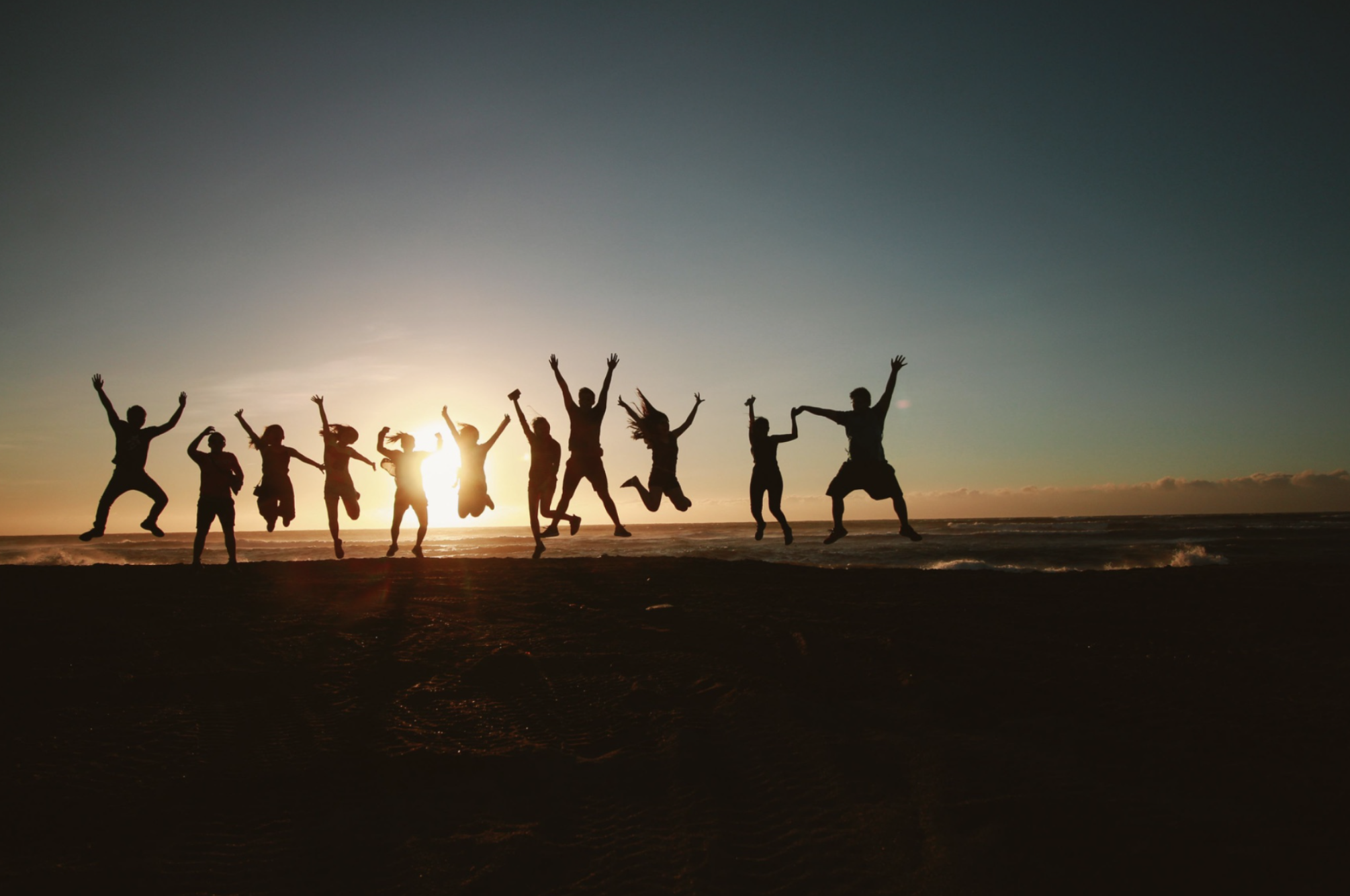 A group of people jumping on the beach at sunset, silhouetted against the sky.