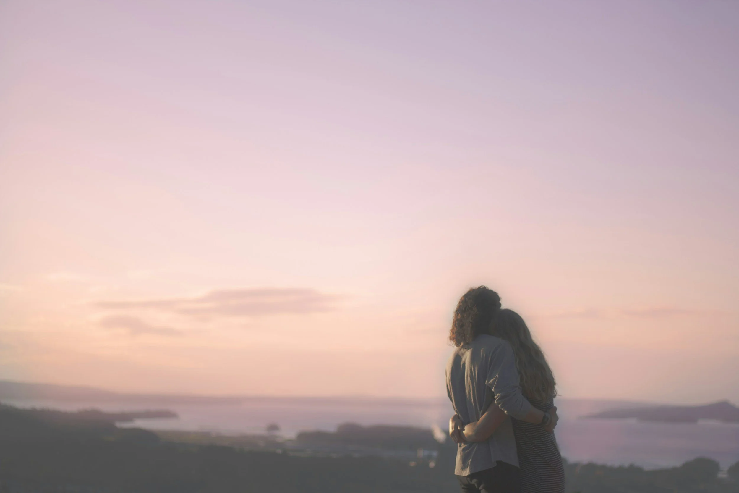 A person with long hair and a striped shirt standing outdoors during sunset, with a pastel-colored sky in shades of pink, purple, and orange, and a landscape of water and hills in the background.
