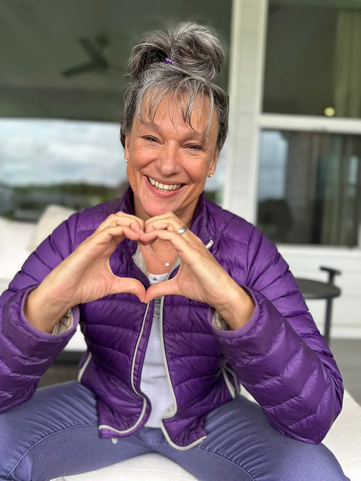 A smiling Rita Henry with gray hair in a bun, wearing a purple puffer jacket, sitting cross-legged outdoors, making a heart shape with her hands.