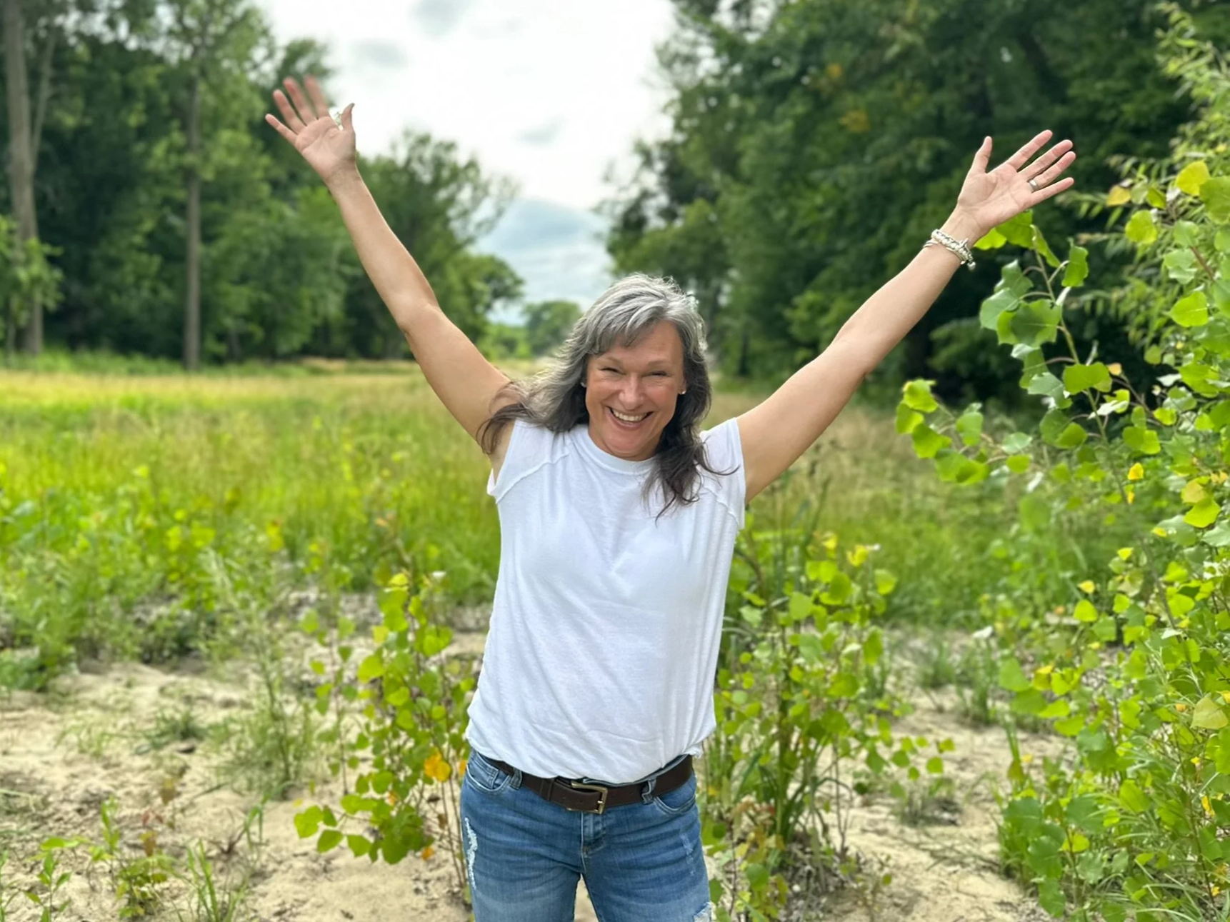 A smiling Rita Henry with gray hair wearing a sleeveless white top and blue jeans, standing in a green field with trees and bushes, raising her arms in celebration.