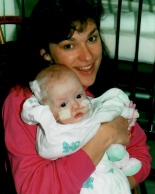 Rita Henry holding a young child with medical tubes attached, inside a hospital or clinic.