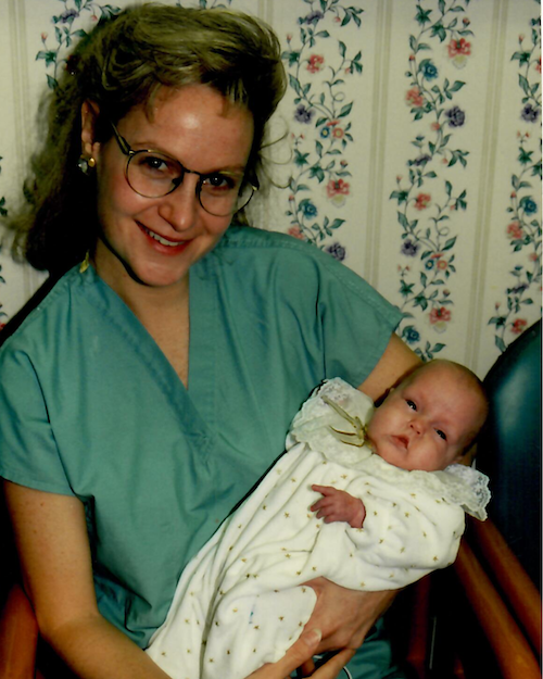 Dr. Teri Wahlig in medical scrubs holding baby Julia Henry indoors, with floral wallpaper in the background.