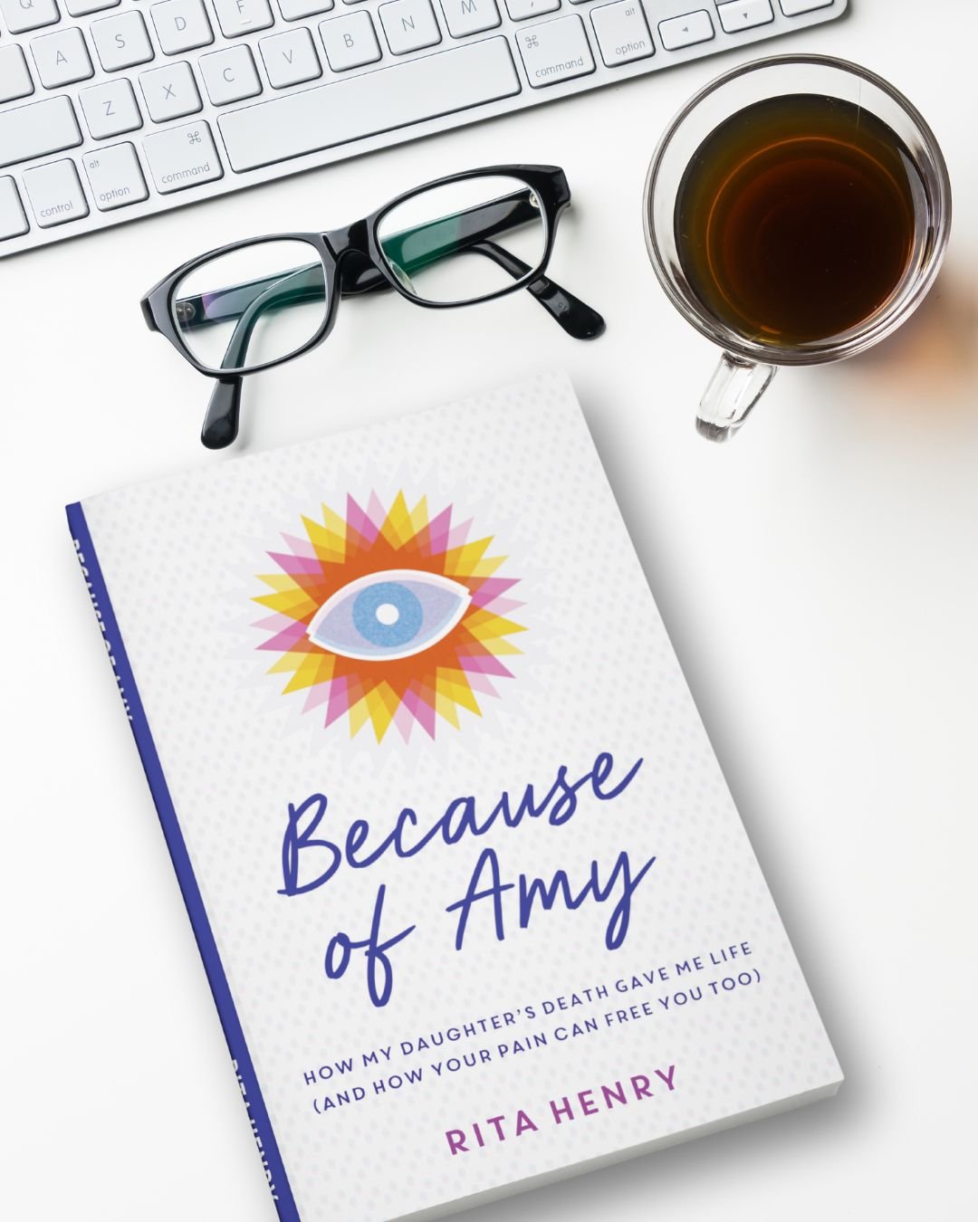 A white desk with a keyboard, a pair of black glasses, a clear mug filled with dark coffee, and a book titled 'Because of Amy' by Rita Henry with a colorful eye illustration on the cover.
