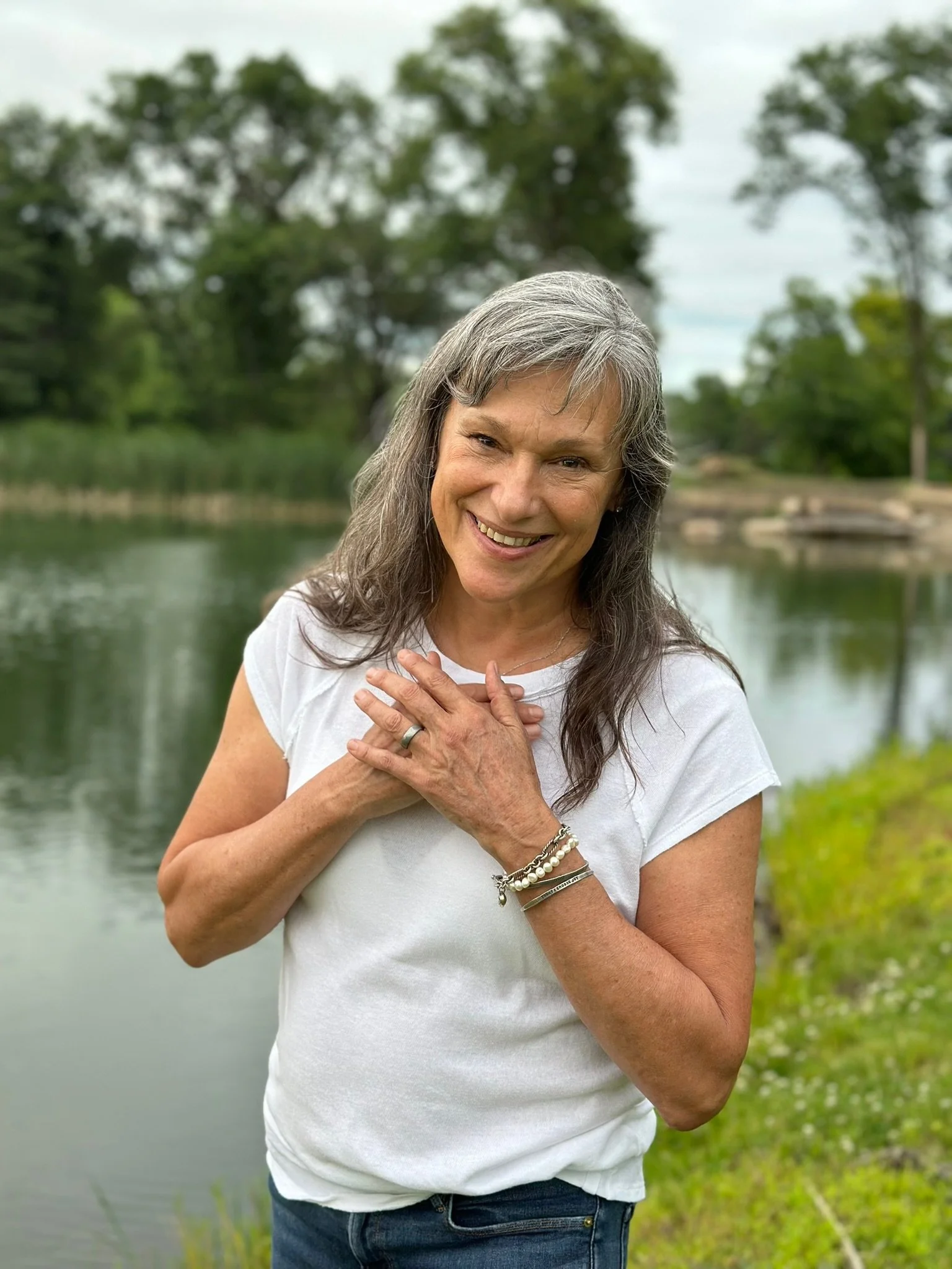 Rita Henry in a white t-shirt, smiling and holding her hands over her heart, standing outdoors near a body of water with trees in the background.