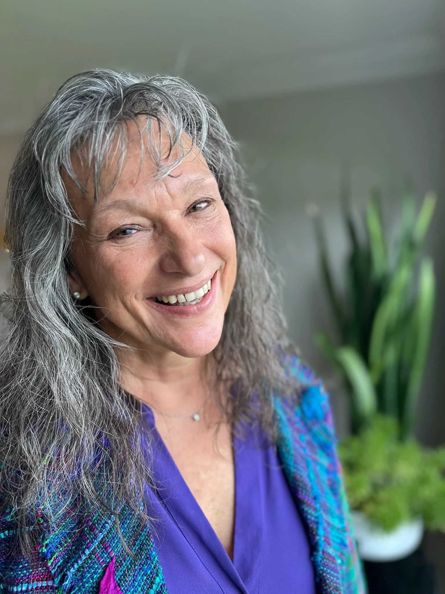 Close-up of a smiling Rita Henry with long gray hair wearing a blue blouse and a colorful jacket, standing outdoors with green plants in the background.