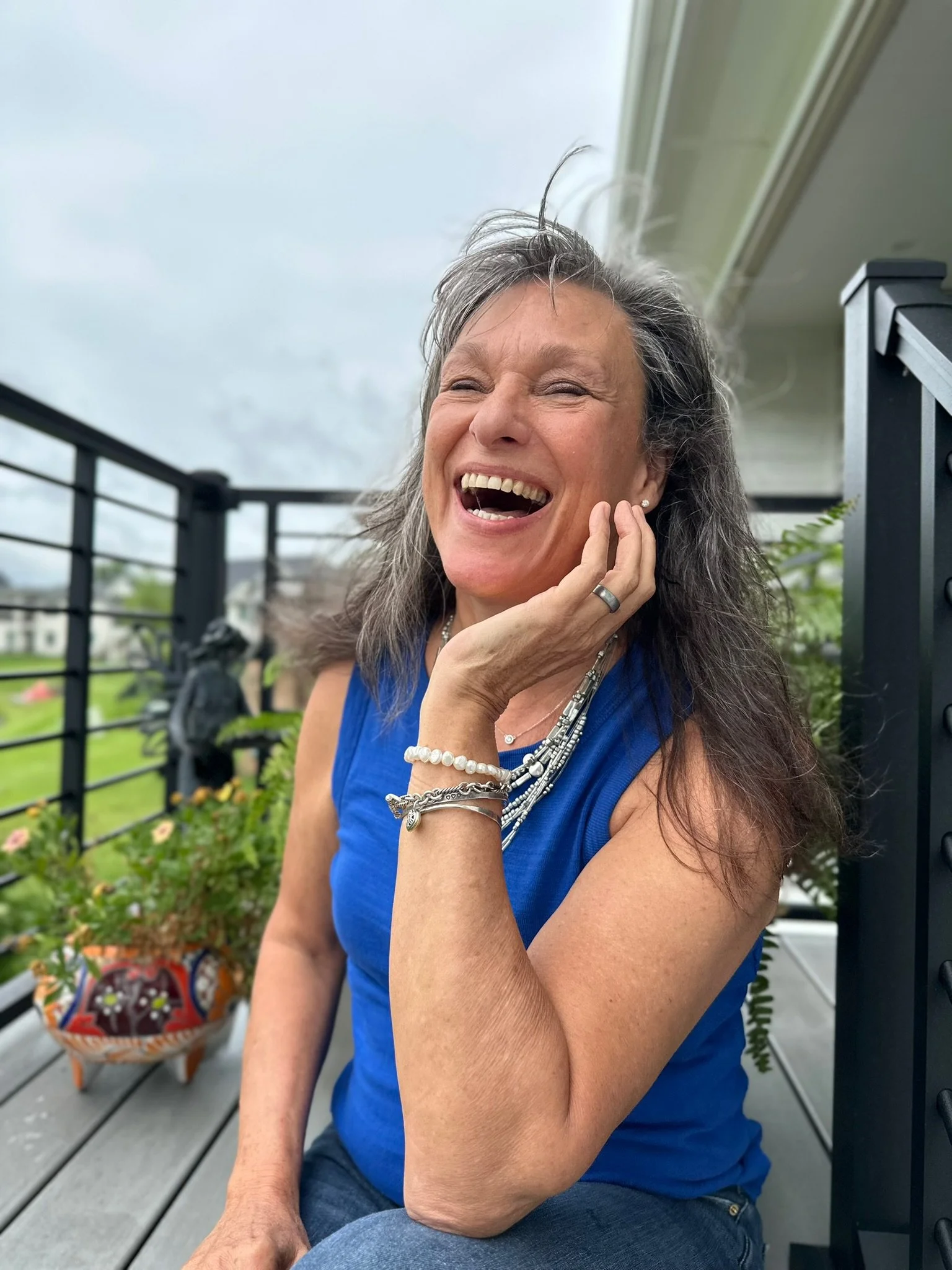 Rita Henry in a blue sleeveless top sitting outdoors on a wooden deck, smiling and laughing with her hand touching her face, surrounded by plants and a black metal railing.