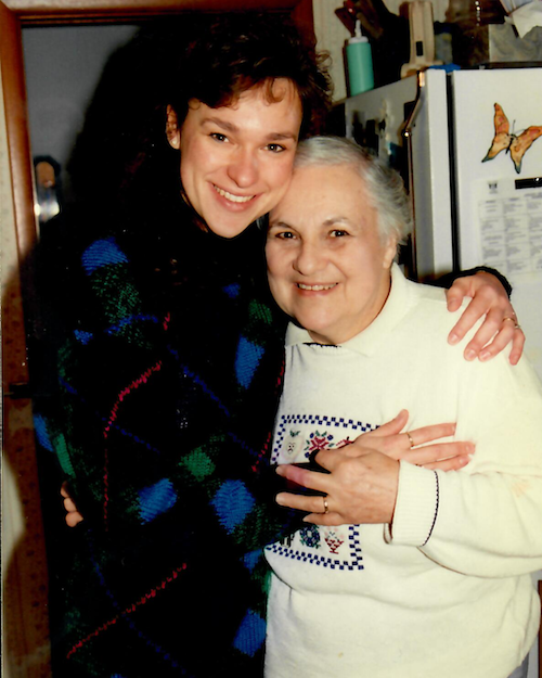 Rita Henry and her mom smiling and hugging in a kitchen. One appears to be younger with dark, curly hair wearing a colorful sweater; the other is elderly with gray hair wearing a white sweater with a decorative pattern.