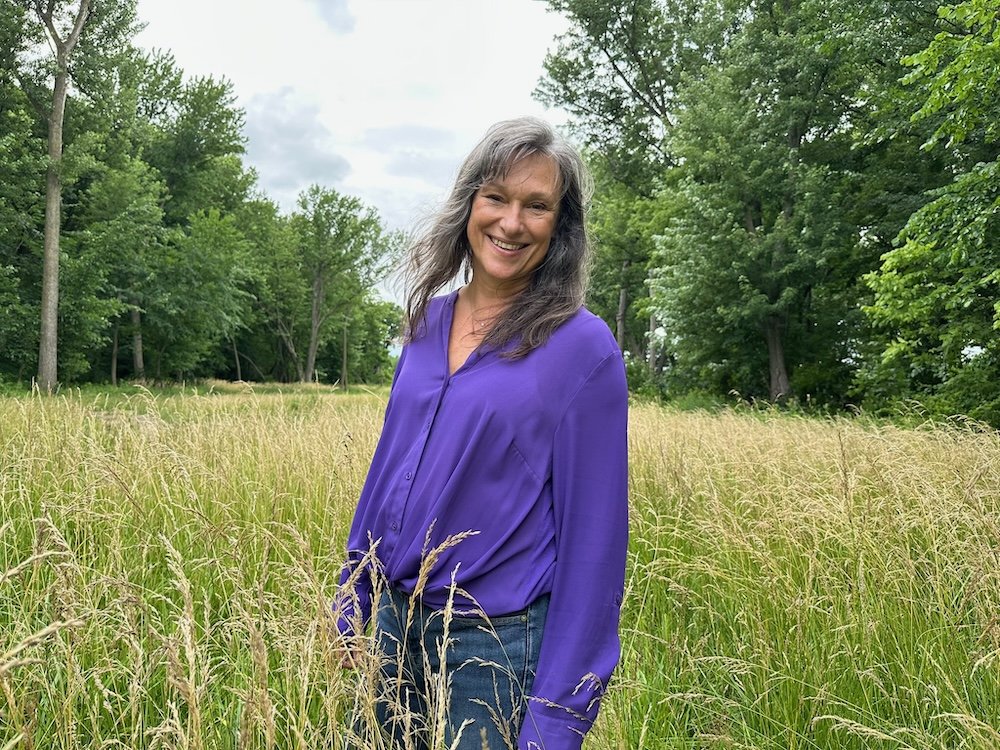 Rita Henry with long gray hair wearing a purple blouse and jeans standing in a grassy field with tall grass and green trees in the background, smiling at the camera.