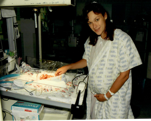 Rita Henry in hospital gown standing next to her newborn baby in a hospital nursery.