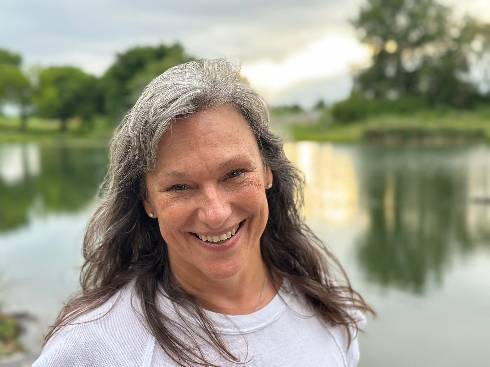 A smiling Rita Henry standing outdoors near a body of water with trees in the background.