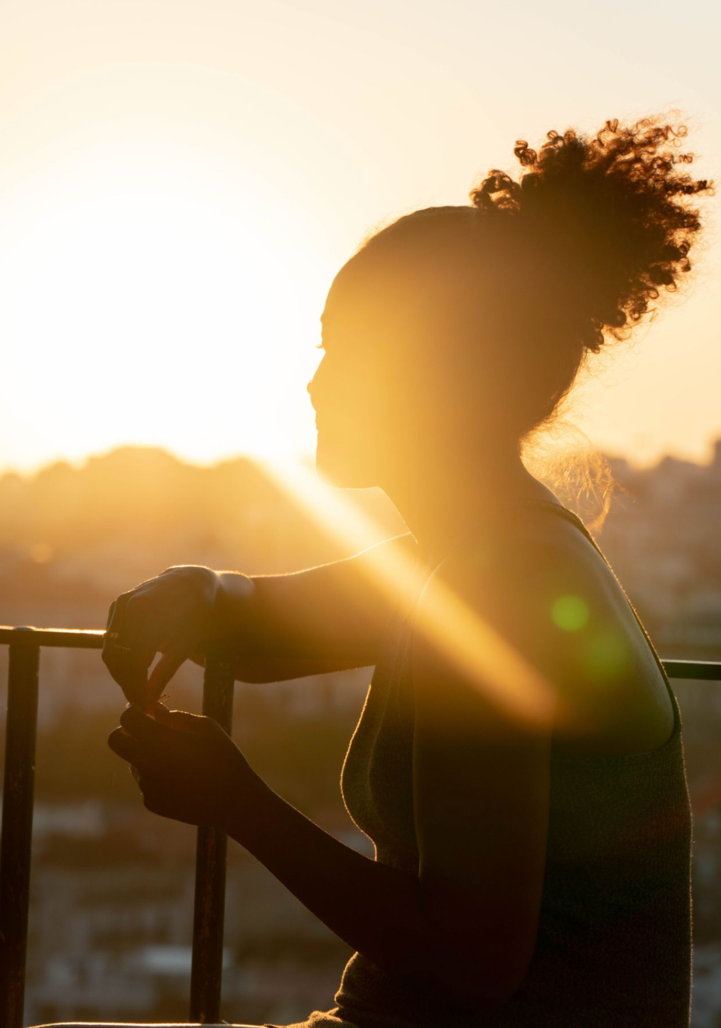 Silhouette of a woman with curly hair tied up in a bun, standing on a balcony during sunset, looking thoughtful.