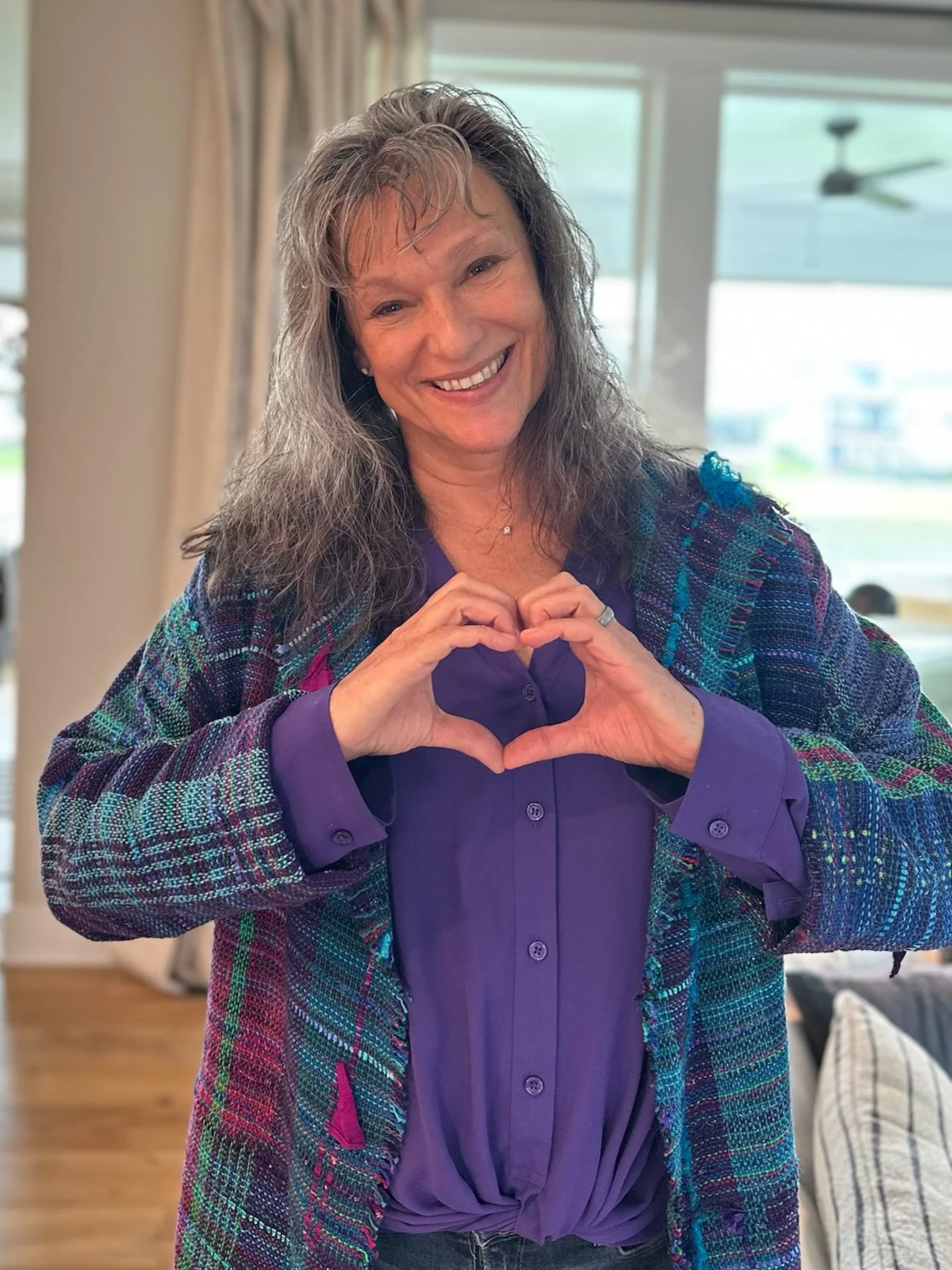 Rita Henry sitting cross-legged on a furry blanket on a couch, smiling with hands on her cheeks, wearing a black jacket and purple striped leggings.