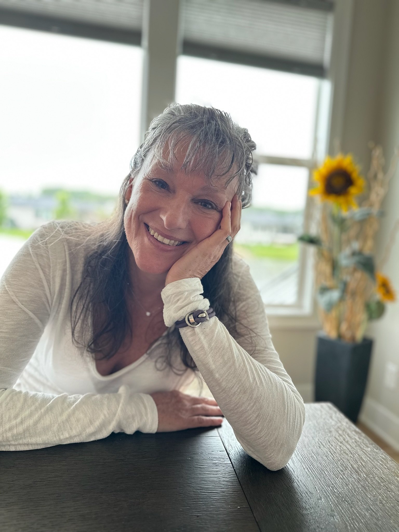 Rita Henry smiling and resting her head on her hand at a dark wooden table in a room with large windows and a sunflower in a vase in the background.