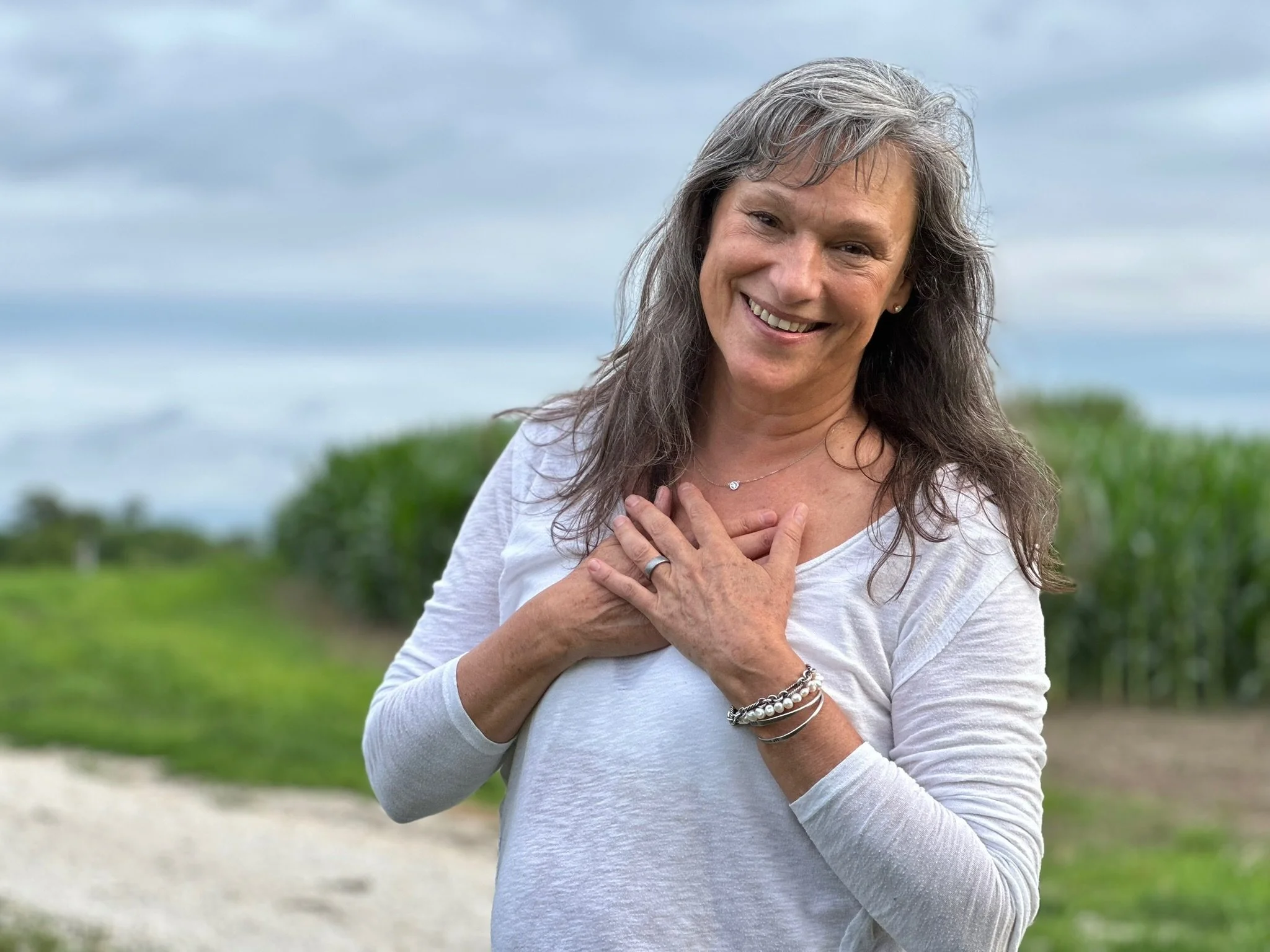 Rita Henry with gray hair standing outdoors, wearing a white long-sleeve shirt, with her hands over her chest, in a green field under cloudy skies.