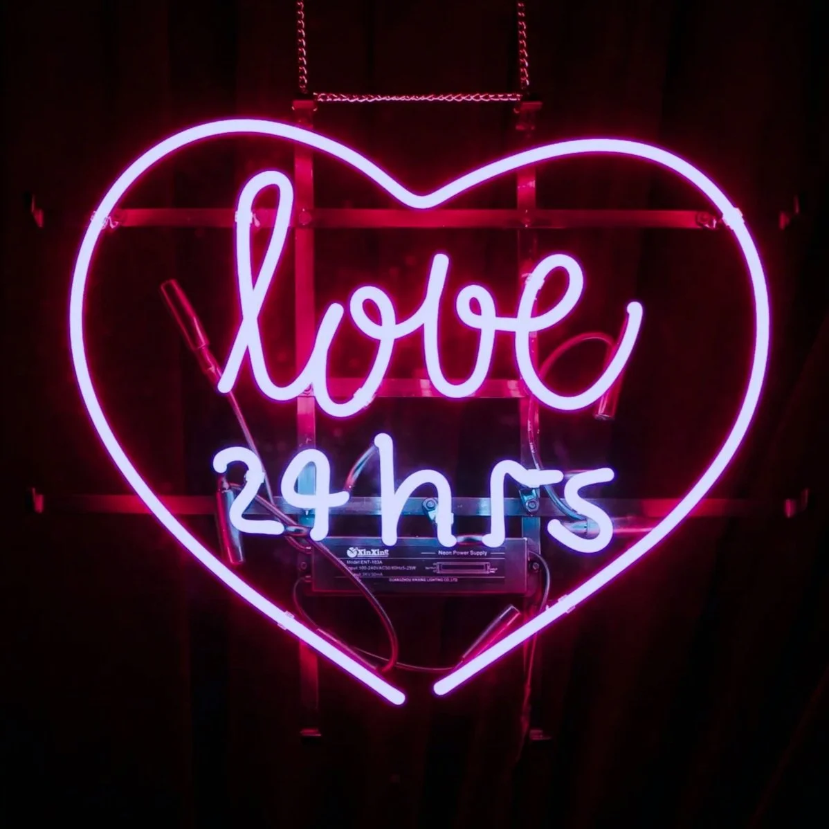 Neon sign with a pink heart outline, featuring the words "love 24 hrs" in the center, illuminated against a dark background.