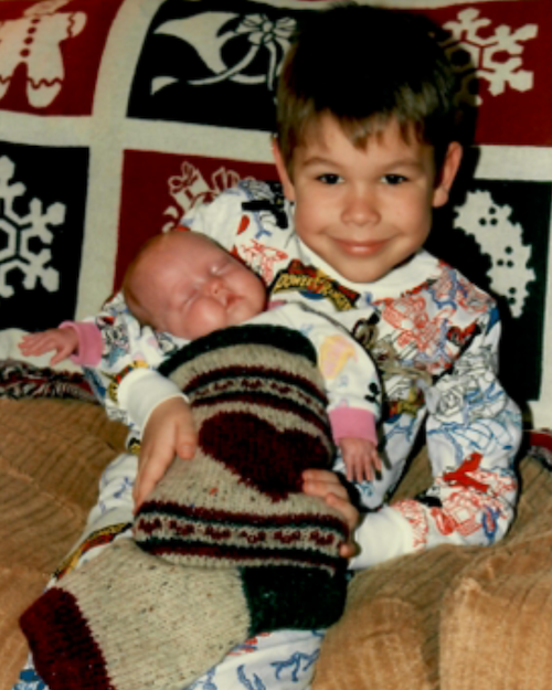 A young boy holding a newborn baby in his arms, sitting on a couch with a festive holiday blanket in the background.