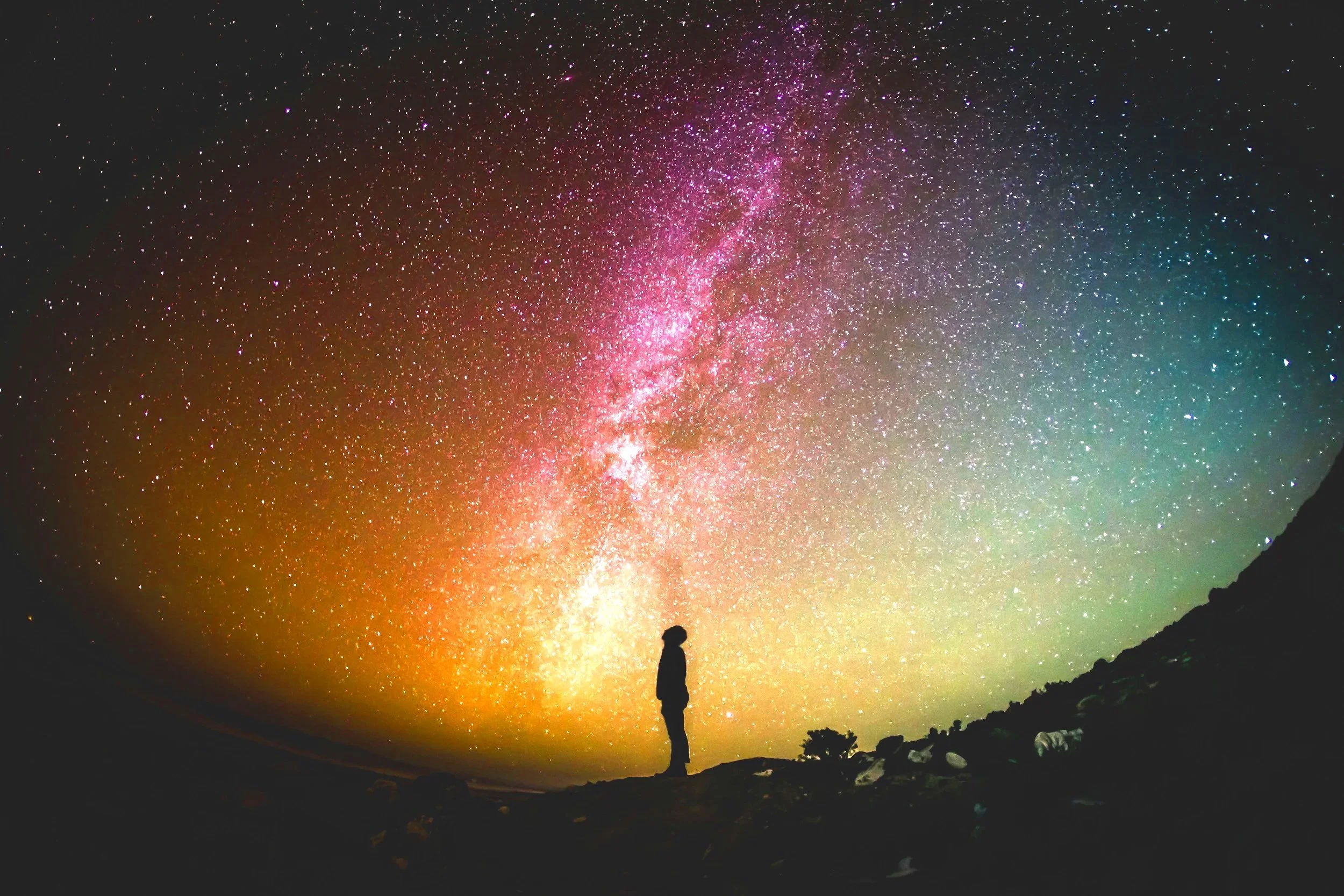 Person standing on a mountain overlooking a colorful, starry night sky with the Milky Way galaxy visible.