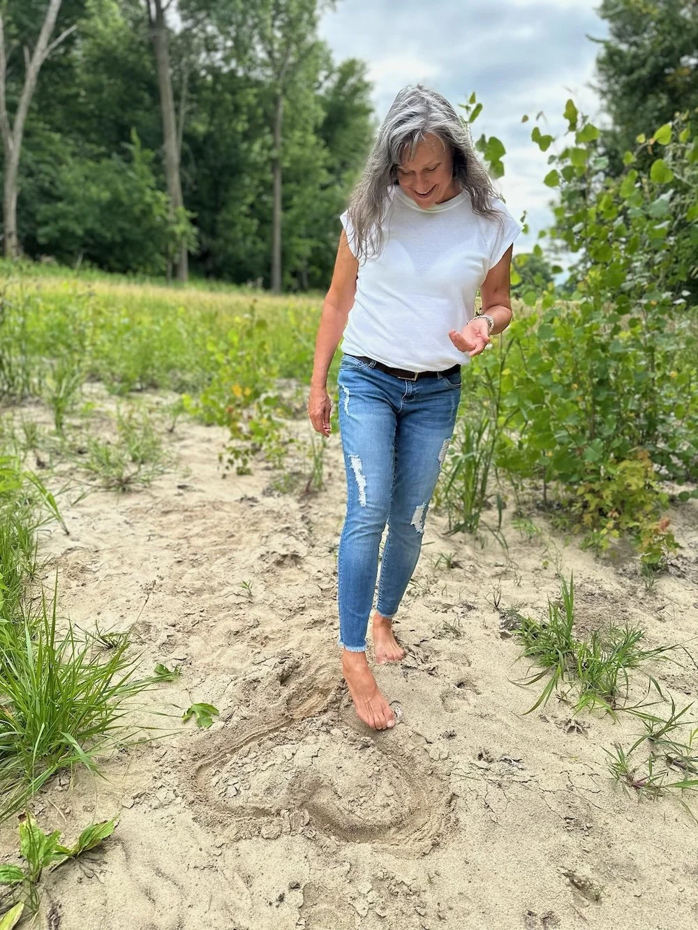 Rita Henry, wearing a white t-shirt and ripped jeans, walking barefoot on a sandy trail in a green outdoor area, smiling and looking down at her foot and a heart she's written in the dirt.