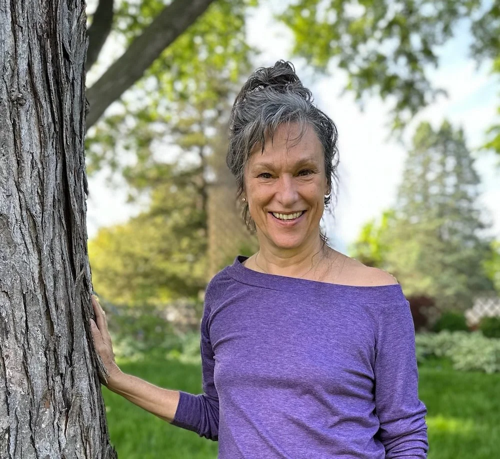 Rita Henry with gray hair in a messy bun, smiling, outdoors with green trees and grass, wearing a purple long-sleeve shirt, standing next to a tree.
