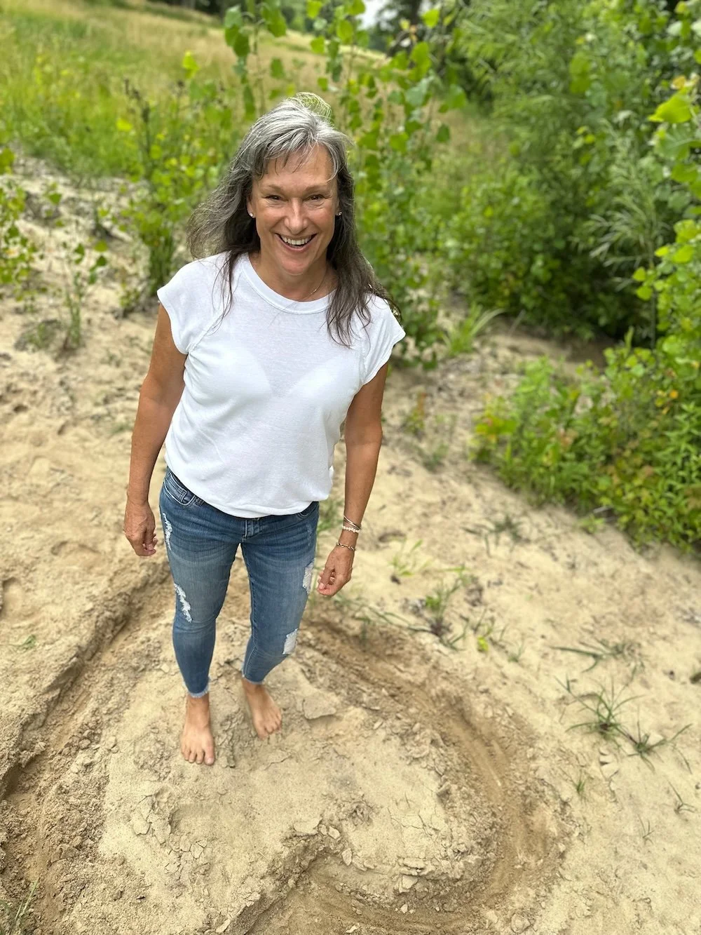 Rita Henry in a white t-shirt and ripped jeans standing barefoot on sandy ground in the middle of a heart, smiling in an outdoor area with green bushes and trees.