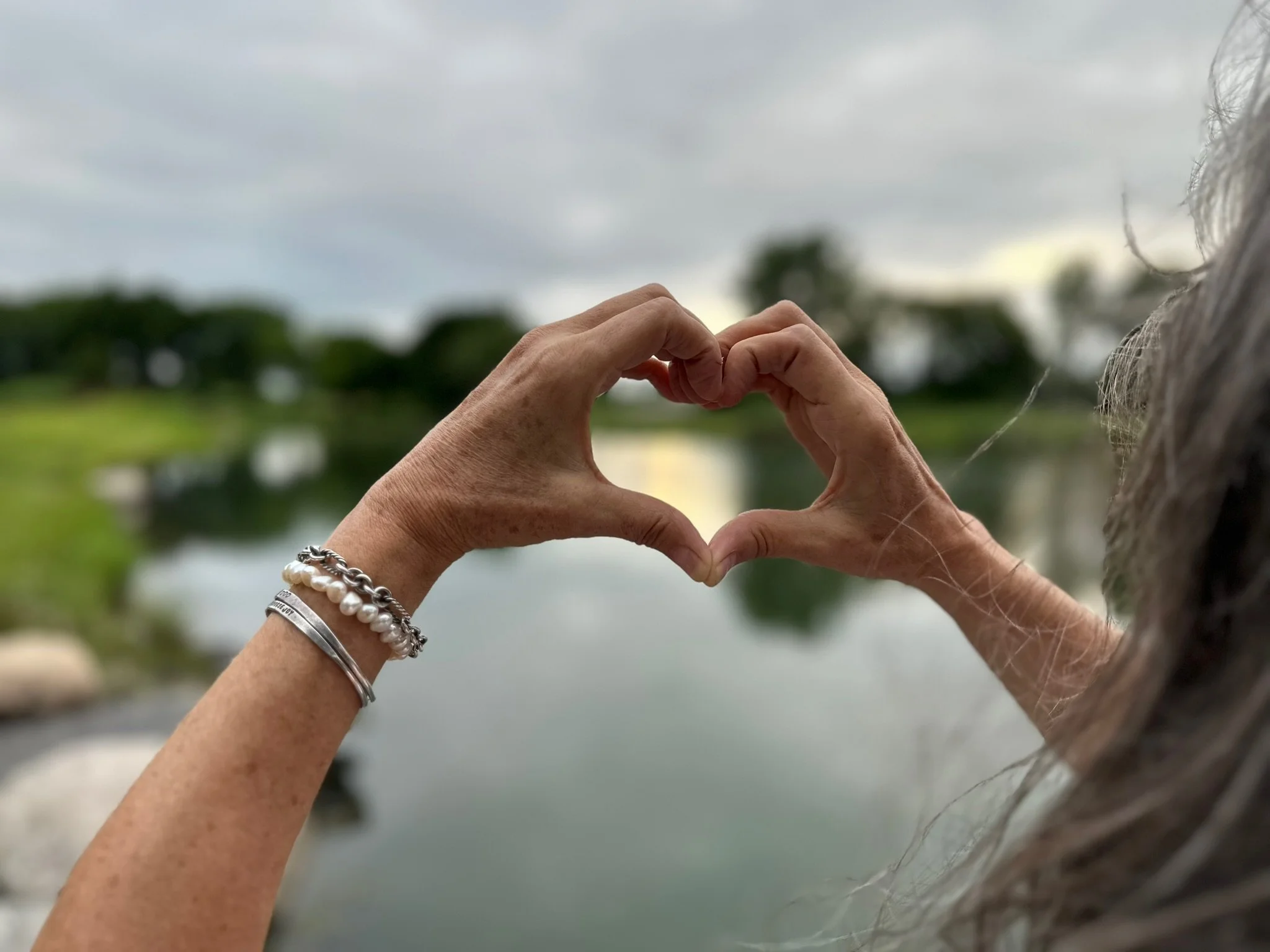 Two hands form a heart shape outdoors, with gray cloudy sky and blurred green trees and water in the background, one wrist with bracelets and the other with a silver bracelet.