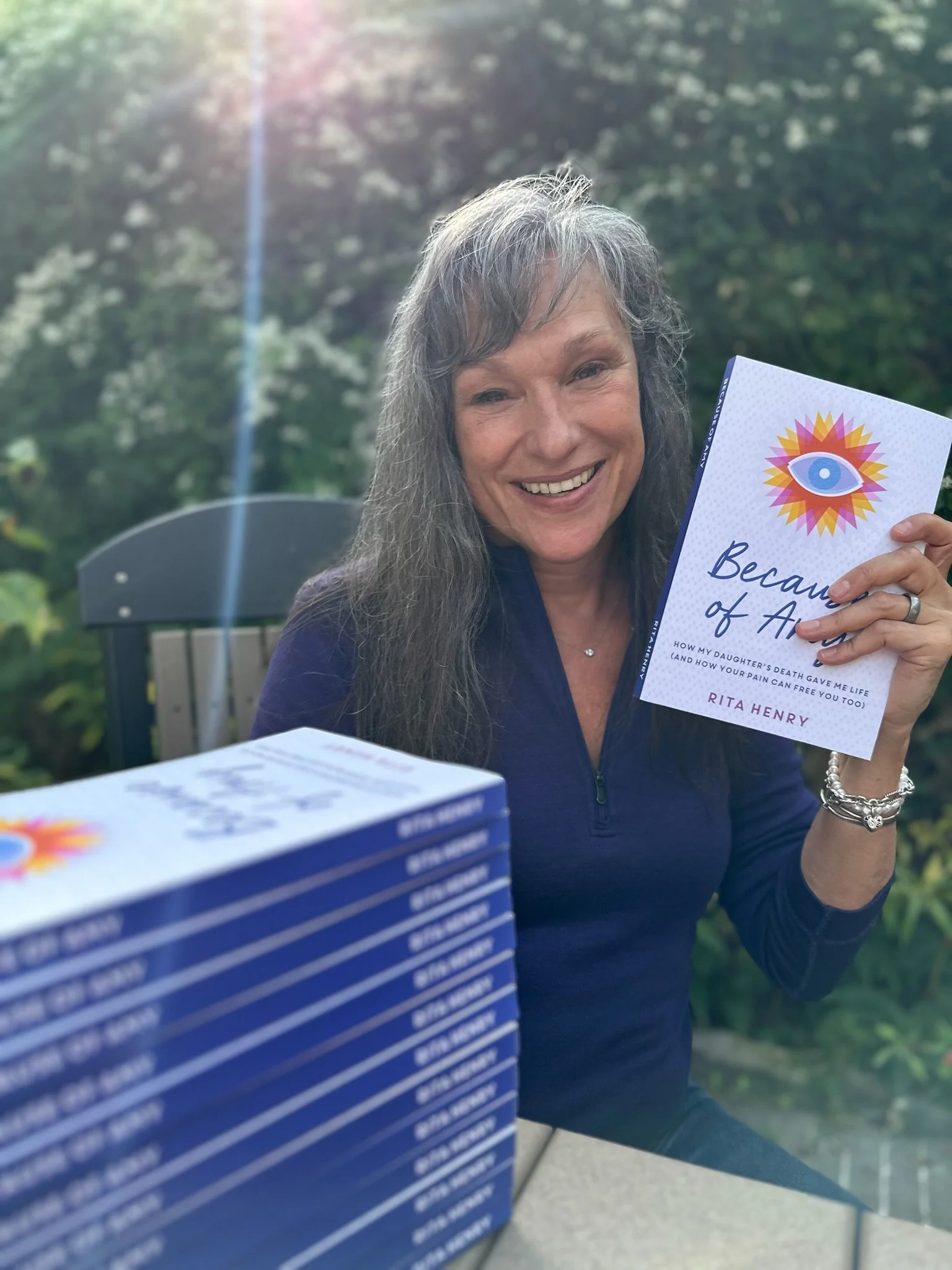 Rita Henry smiling outdoors while holding a book titled 'Because of Angel' by Rita Henry, with a background of trees and sunlight.