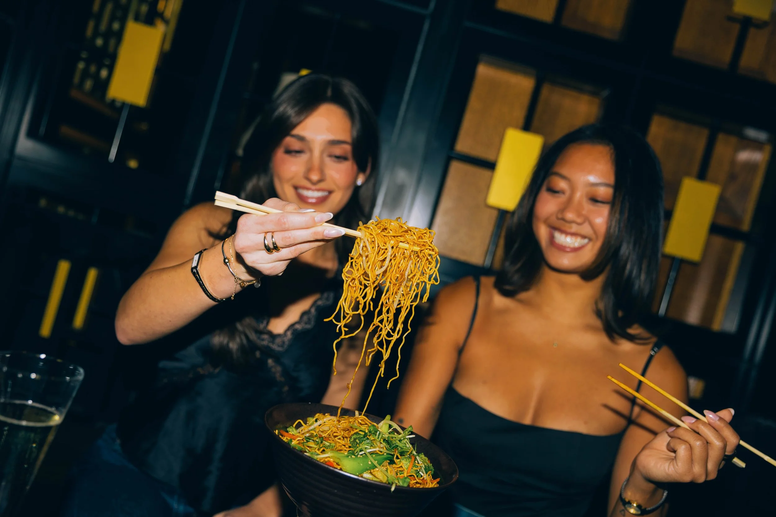 Two women enjoying a meal at a restaurant, one of them holding a bowl of Asian noodles with chopsticks.