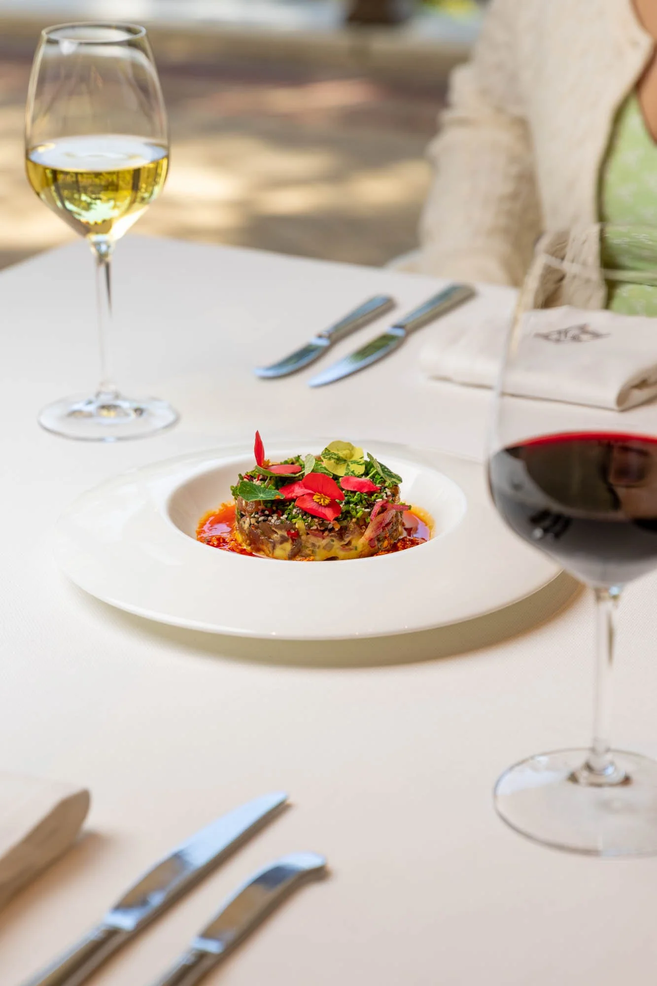 A restaurant table set with a glass of white wine, two forks, and a plate with a gourmet dish garnished with red and green herbs, in front of a person wearing a beige sweater and a green top. food and beverage photography . 
