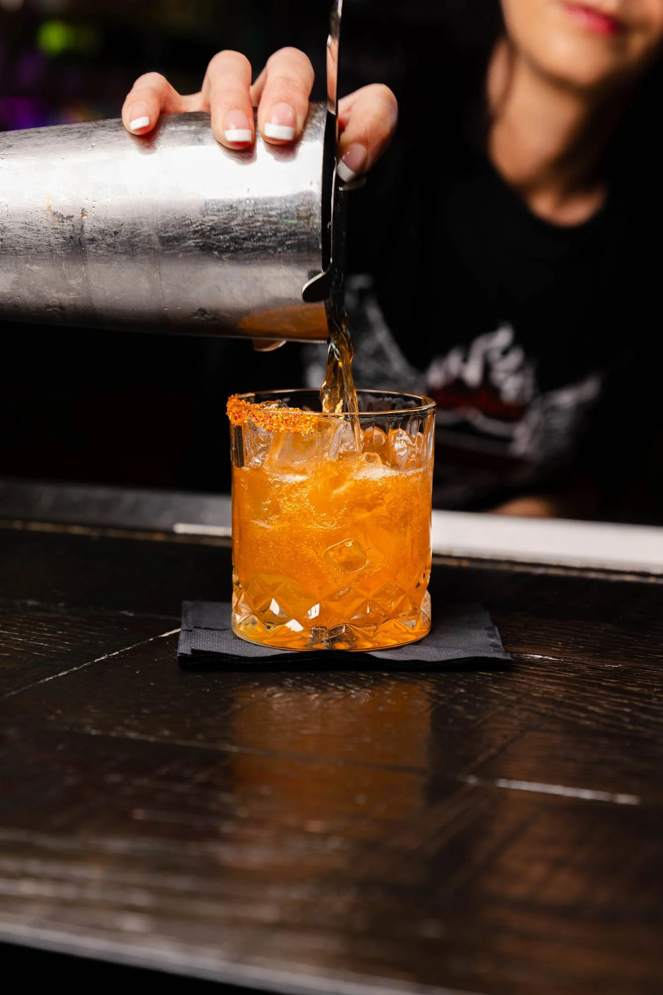 A person pouring a dark-colored drink from a shaker into an orange cocktail glass with ice, garnished with a rim of chili powder or spices, on a dark wooden surface. food and beverage photography.