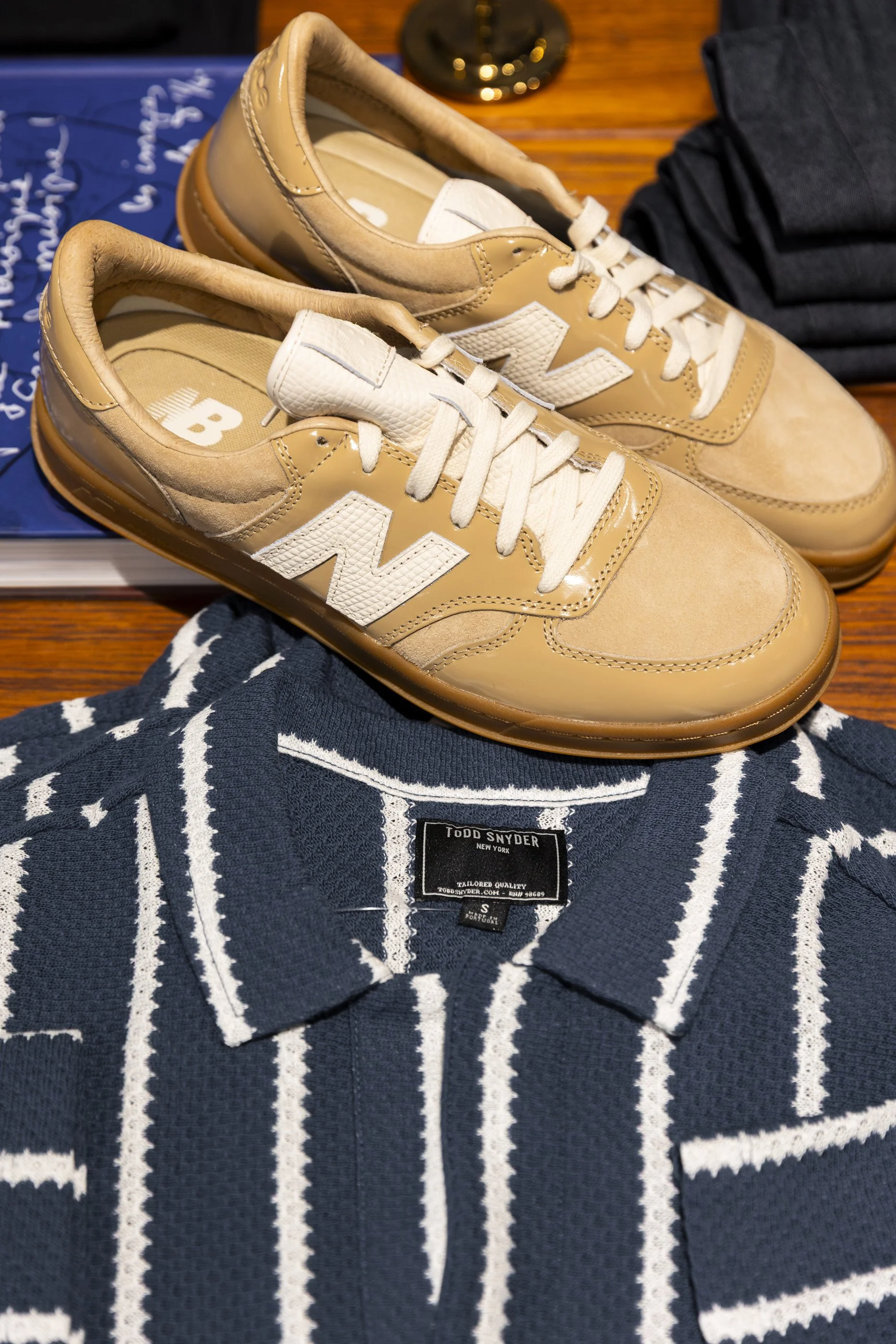 Pair of beige and white sneakers on a wooden table next to a navy and white striped sweater and a blue book with writing. A black bag is also partially visible.