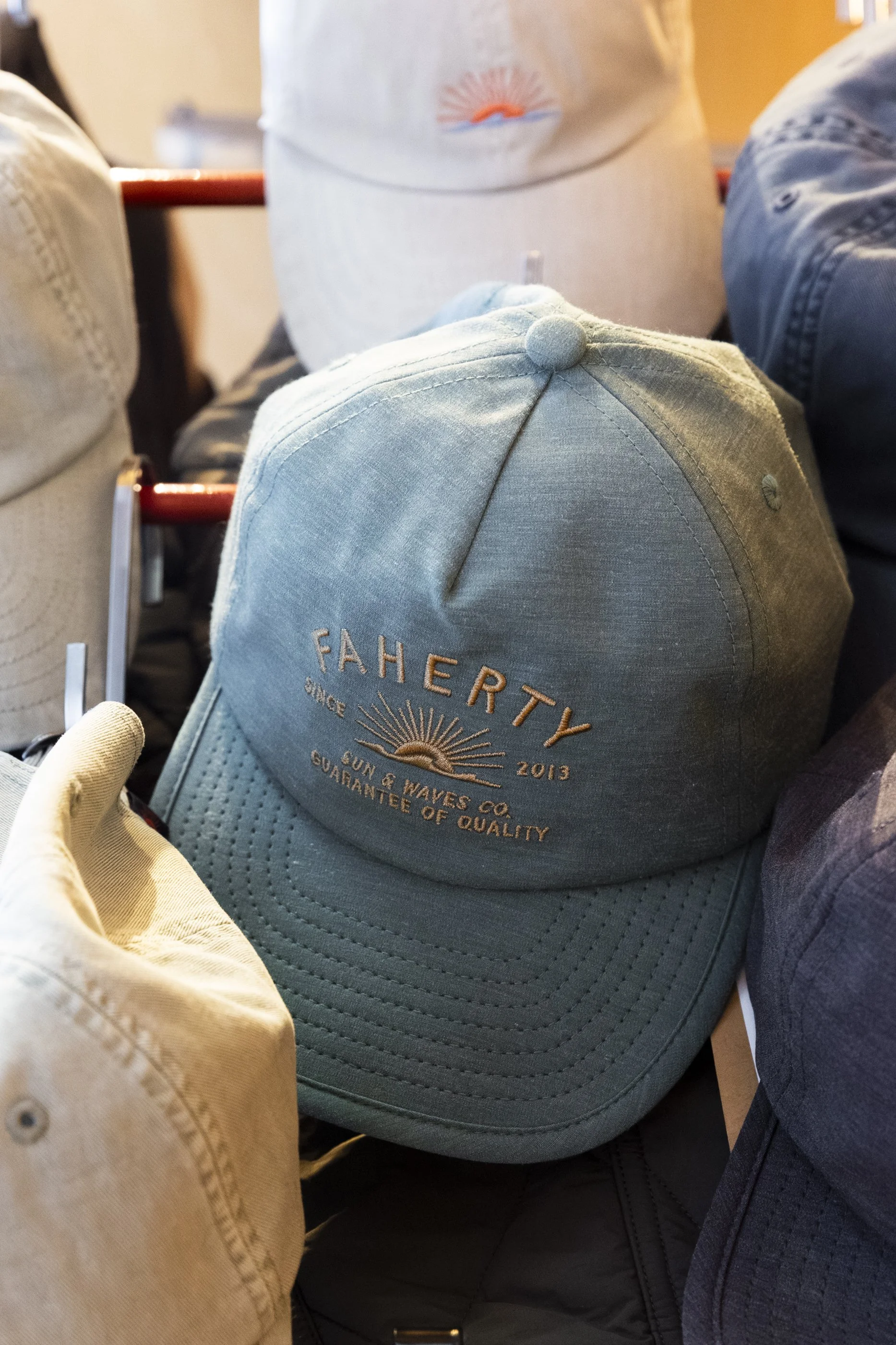 Close-up of a light blue cap with embroidery reading 'FAHERTY' and a sun and waves design, surrounded by other hats and clothing items on a display.