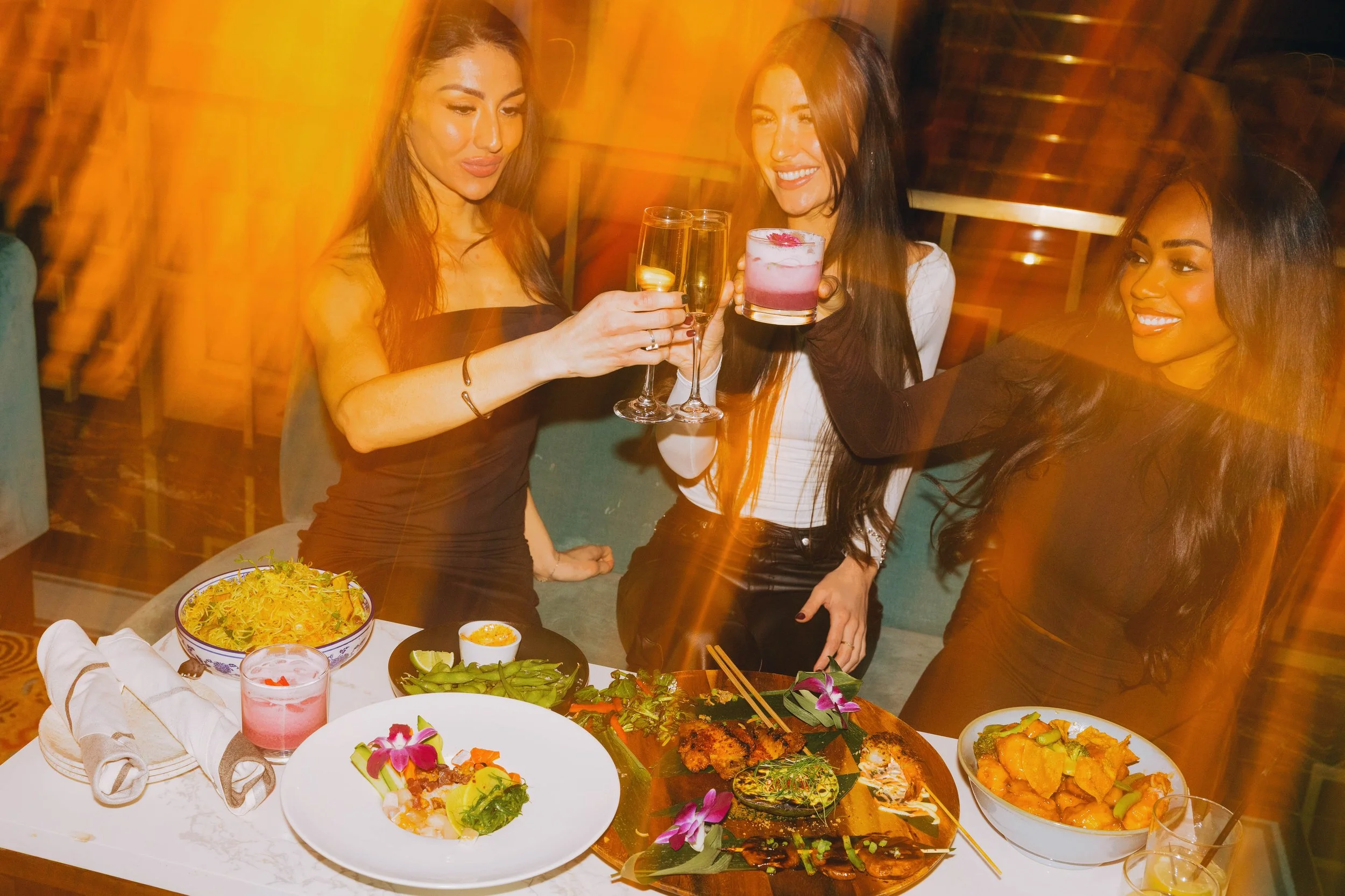 Three women enjoying drinks at a dinner table with various Asian dishes, such as sushi, grilled meats, and vegetables, in a warmly lit restaurant. food and beverage photography.