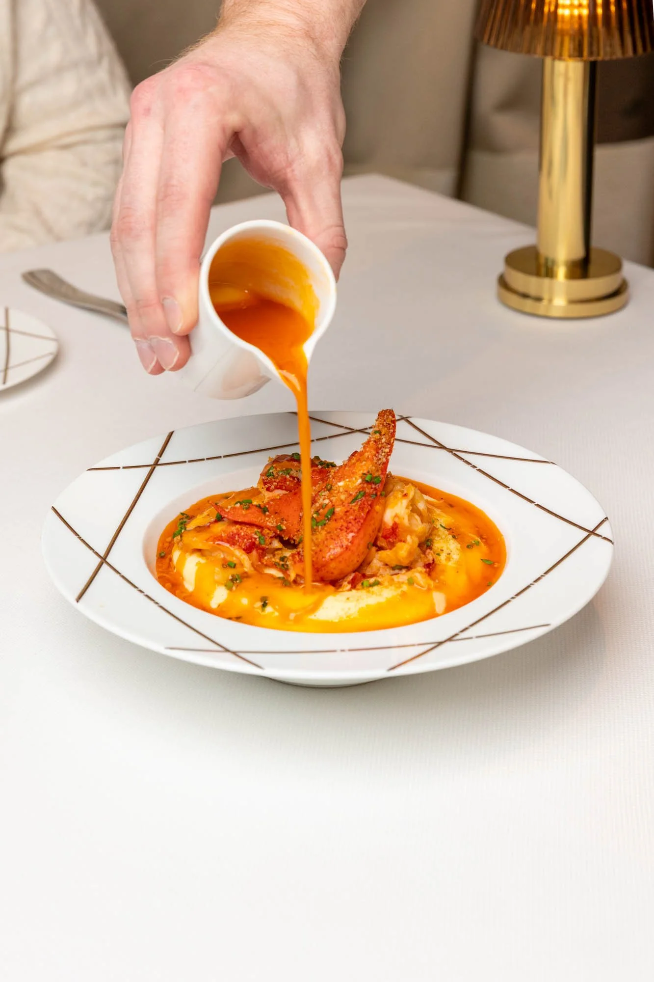 A person pouring orange sauce over a bowl of seafood pasta with lobster tail, garnished with herbs, on a white table with a gold lamp in the background. food and beverage photography