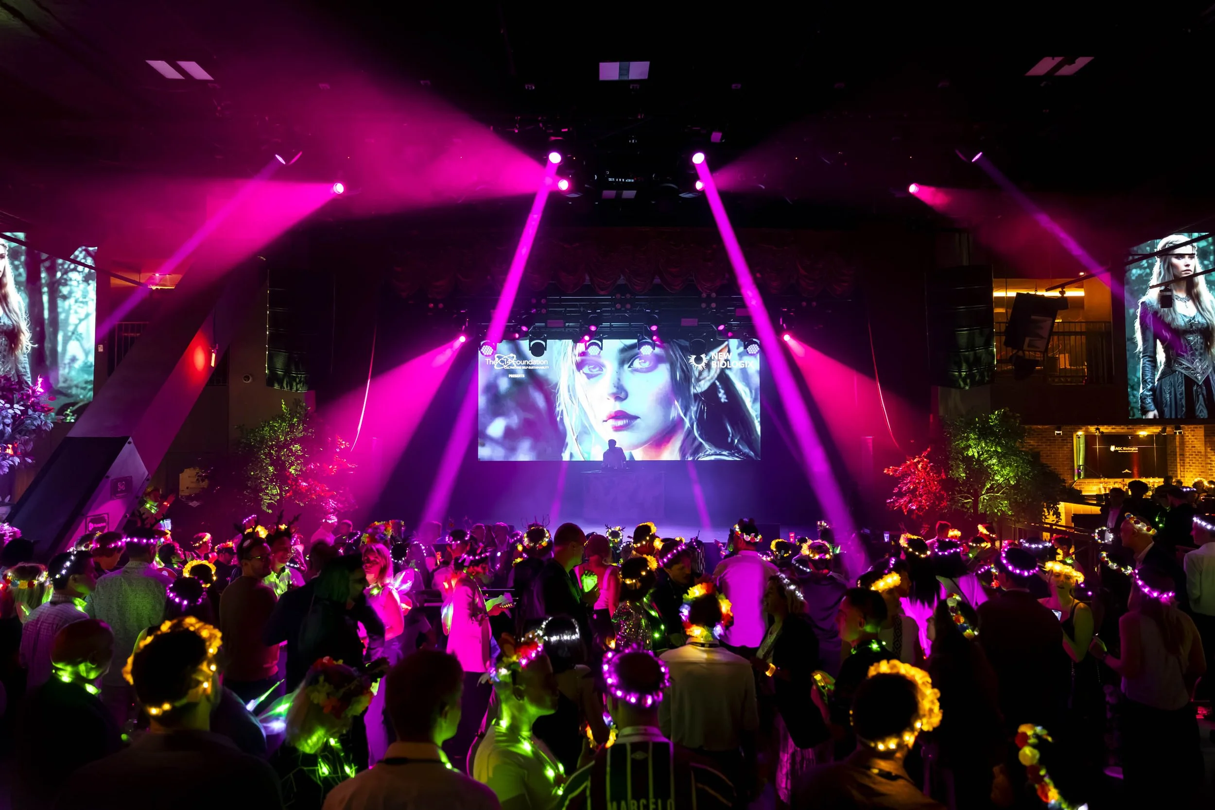 An indoor concert or event with purple and pink stage lighting, a large crowd wearing glowing accessories, and a big screen showing a woman's face. Event concert photography. 