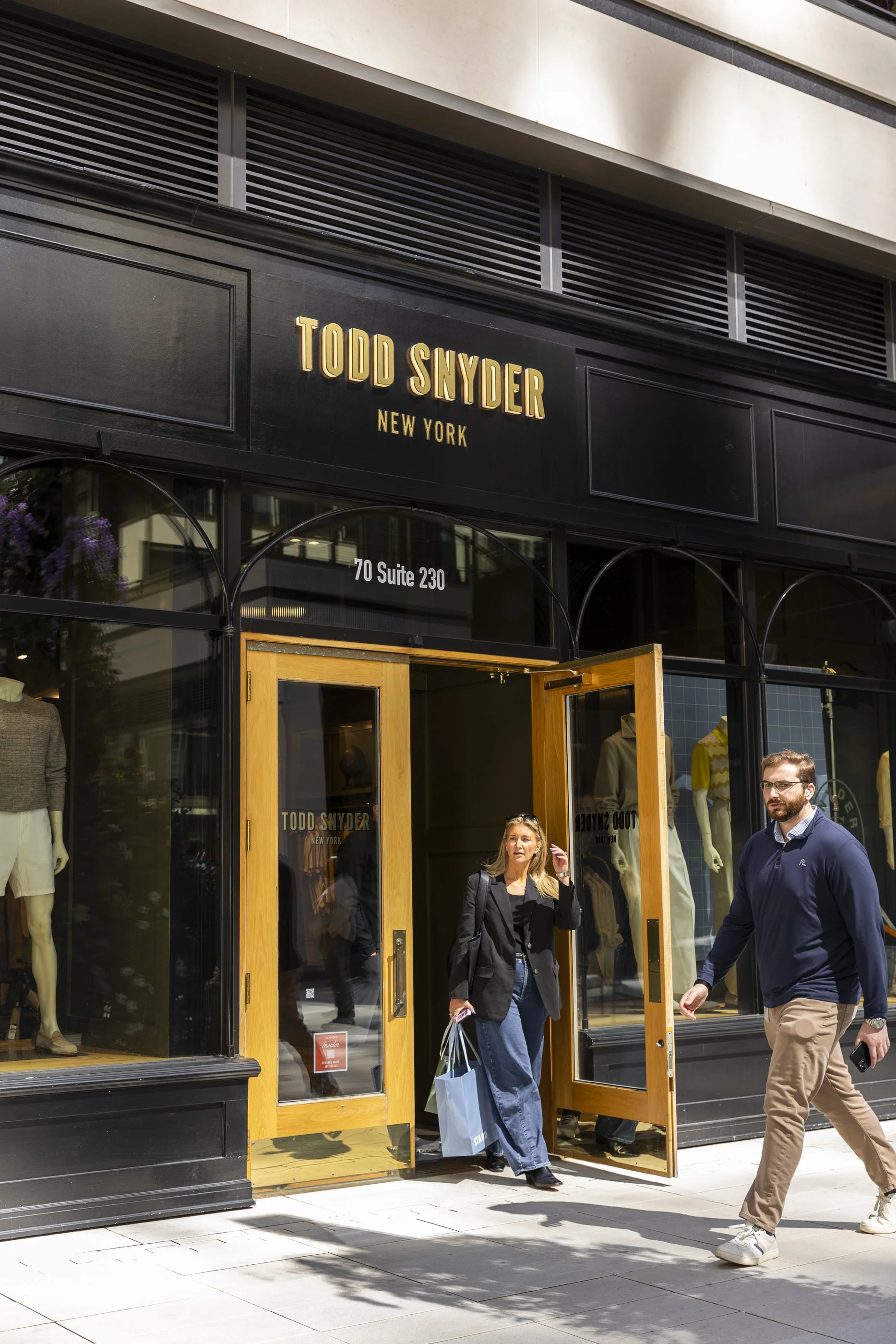 People walking out of a Todd Snyder fashion store on a city sidewalk, with mannequins dressed in clothing visible inside and shopping bags in hand.