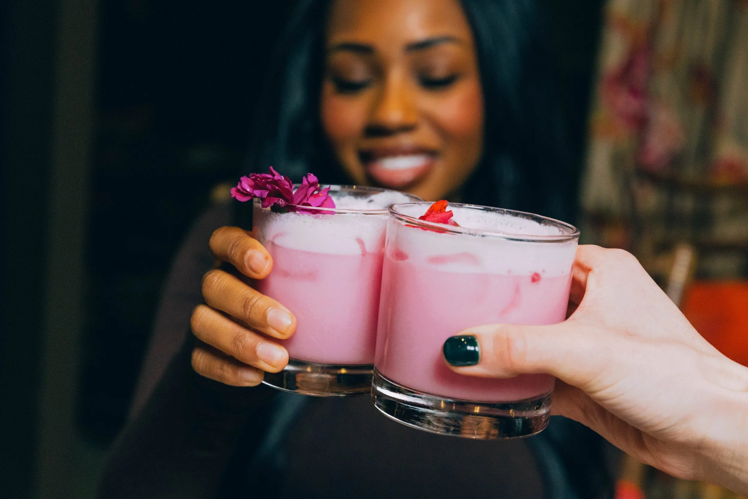 A woman with dark hair smiling while holding two glasses of pink cocktails topped with foam and garnished with flower petals.