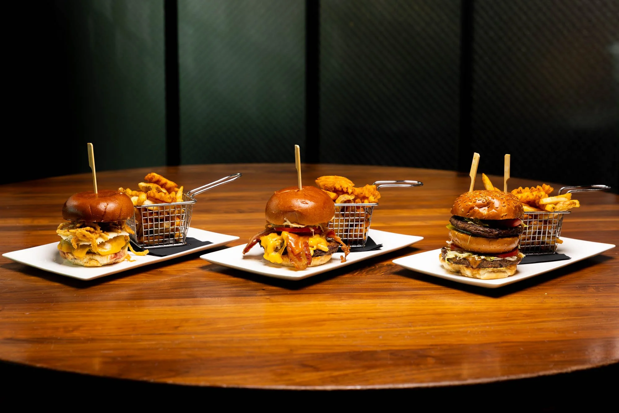 Three plates of burgers and fries on a wooden table, with a dark background. food and beverage photography.
