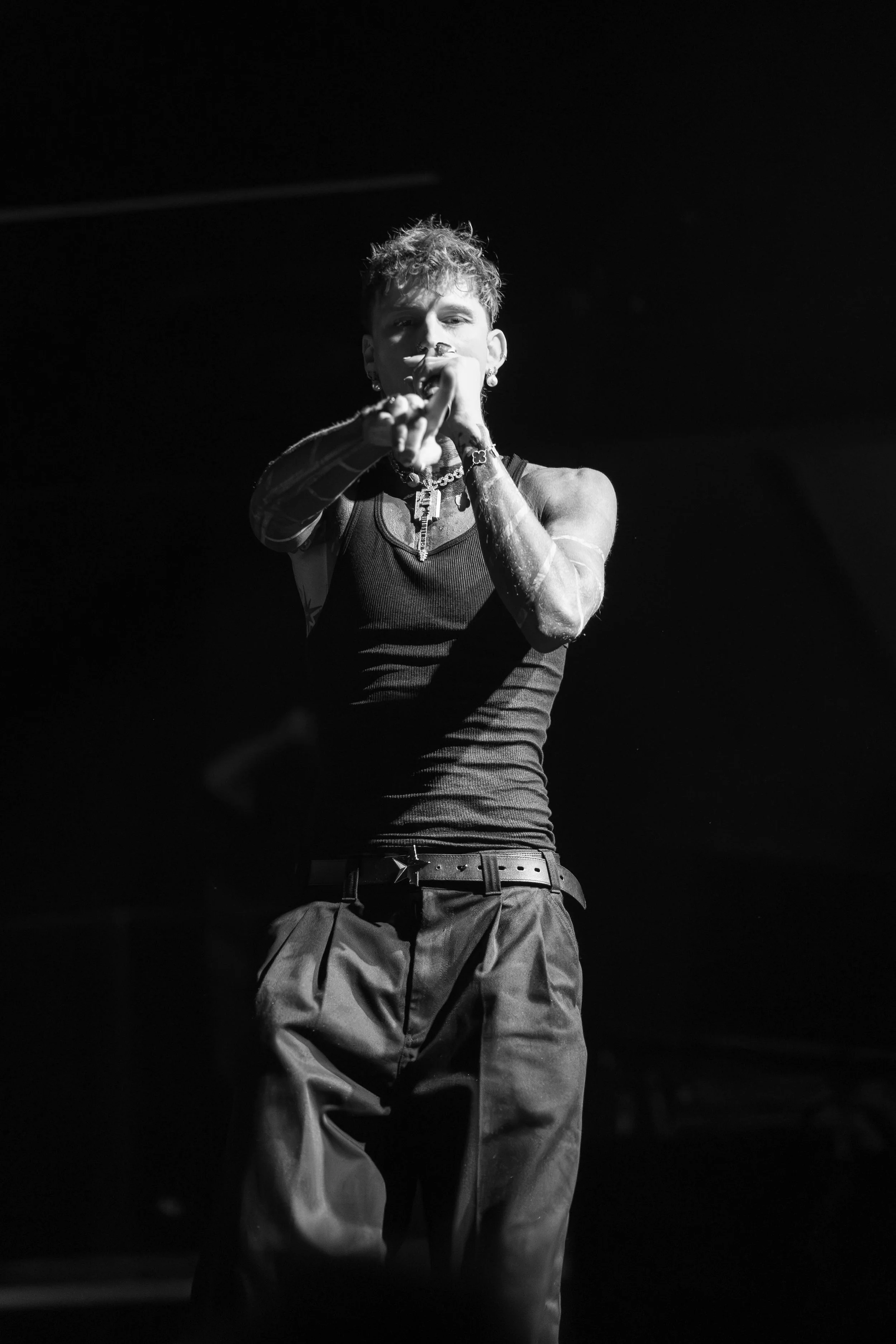 Black and white photo of a young person with curly hair, wearing a tank top and baggy pants, pointing directly at the camera. machine gun kelly nightlife photography. 