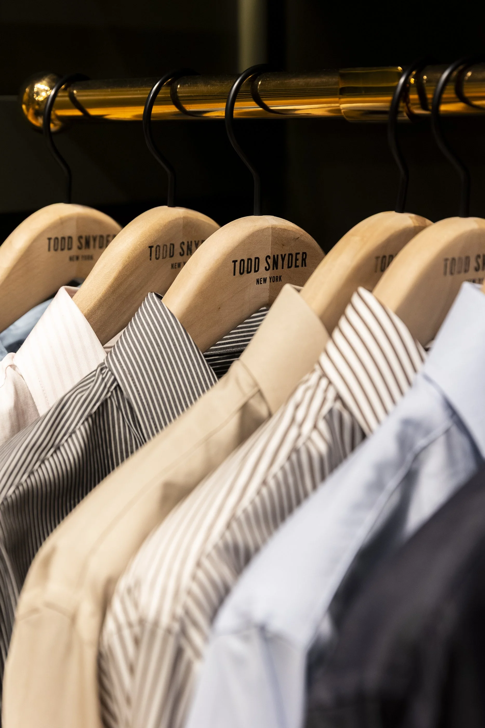 Clothes on wooden hangers with 'Todd Snyder New York' branding, hanging on a gold-colored garment rod.