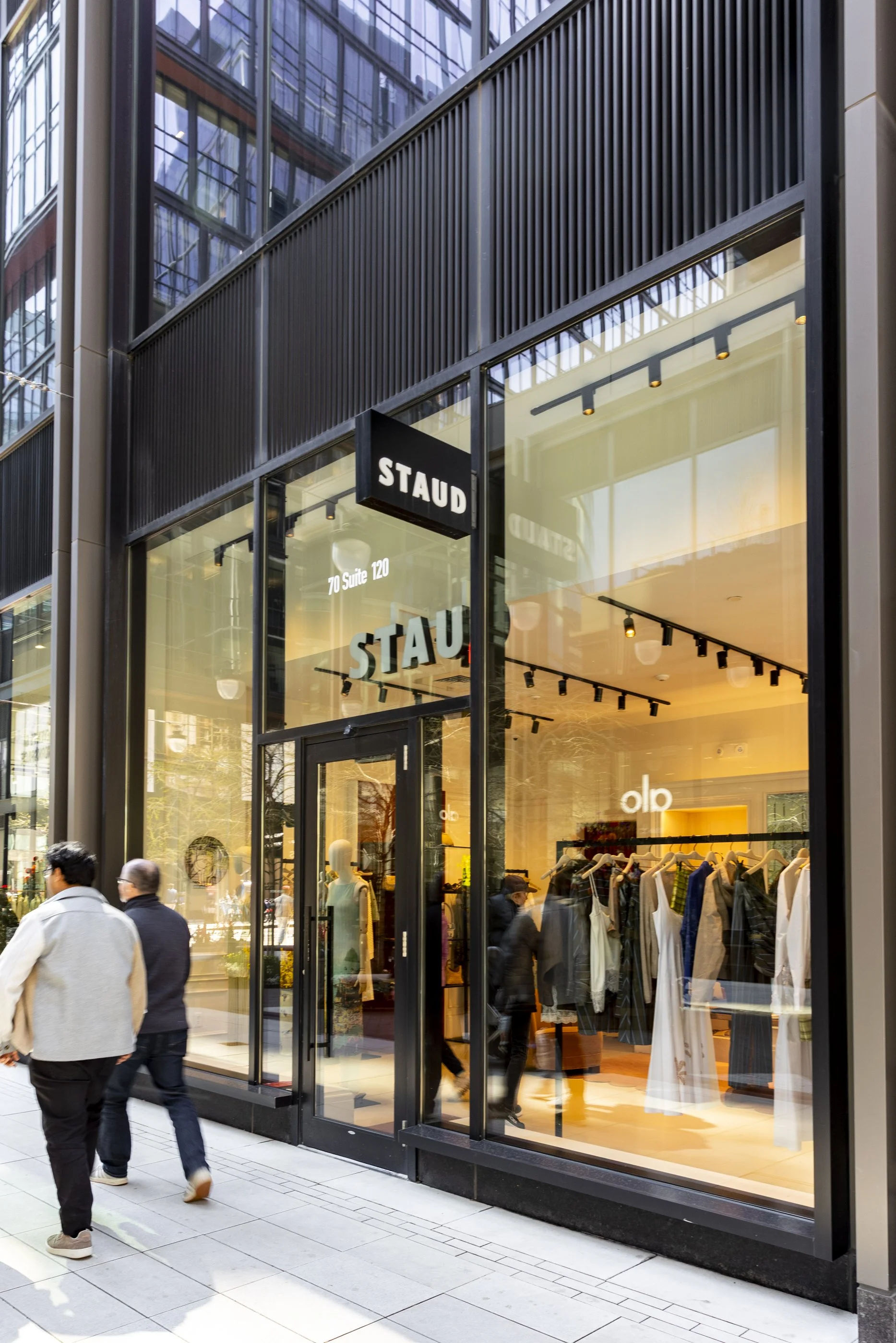 People walking past a storefront with large glass windows displaying clothing inside, and a sign reading 'STAUD' at 70 Suite 120.