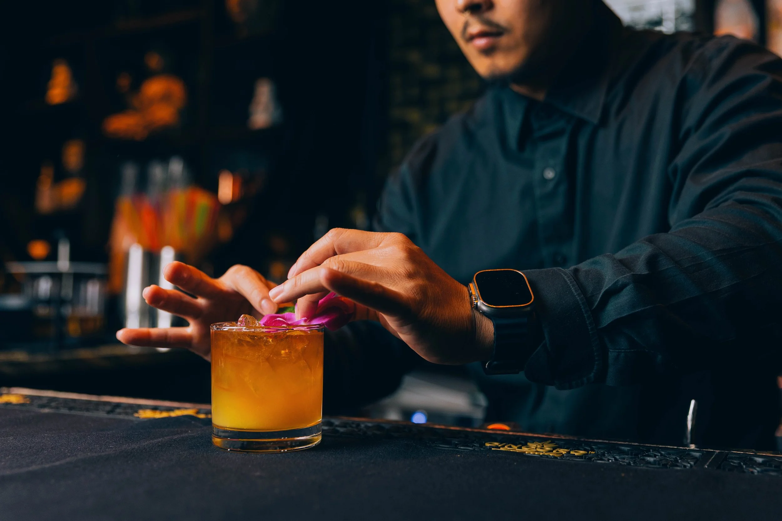 A person wearing a black long-sleeved shirt and a smartwatch, preparing a cocktail with pink flower garnish in a bar or restaurant setting. Jack MacMullin, Boston, Seaport, Photographer, Videographer, Event photography, Event video, Event video cover